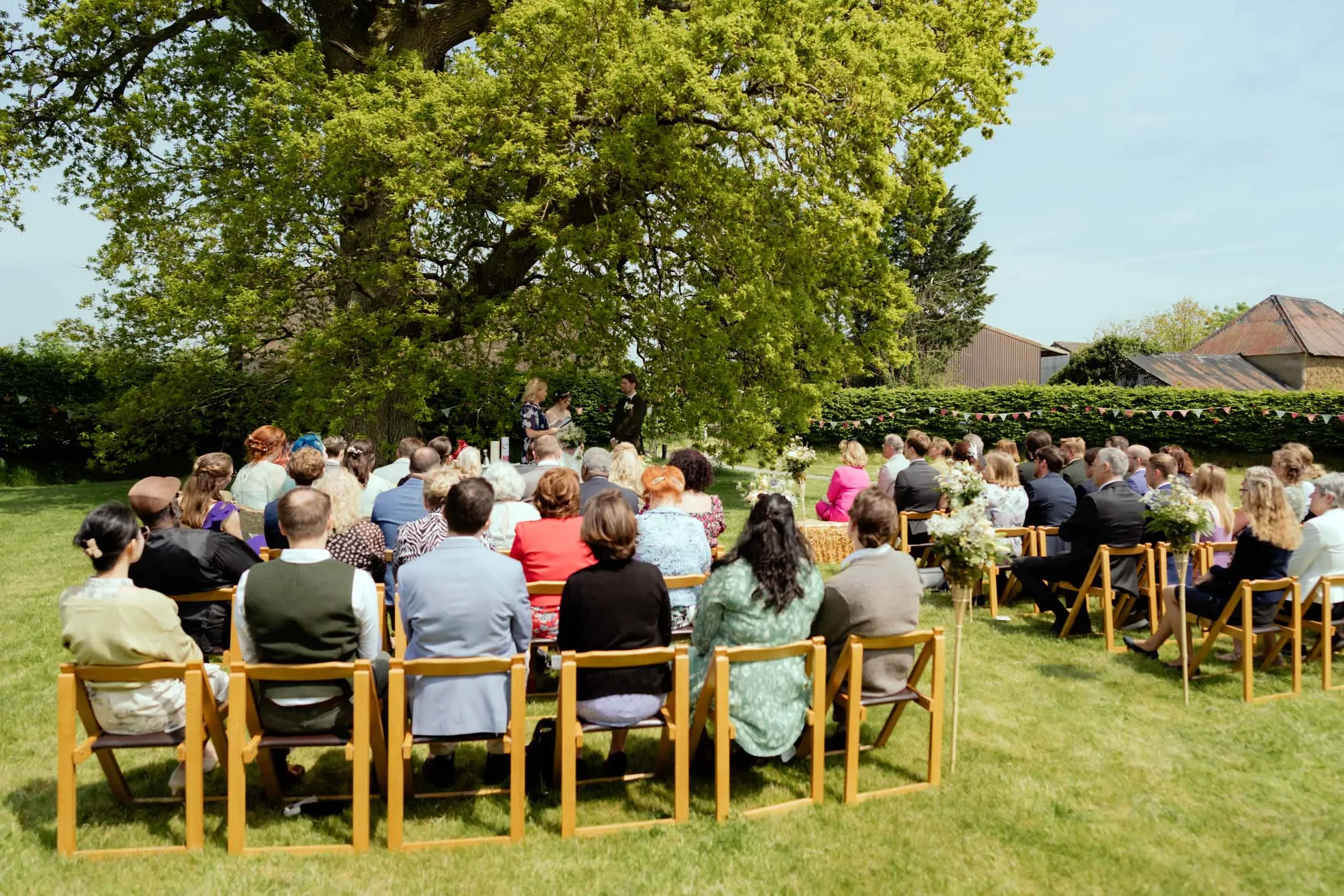 Outdoor wedding ceremony with guests seated on wooden chairs under a large tree, officiant and groom standing in front of wedding couple, sunny day with green grass and buildings in background.