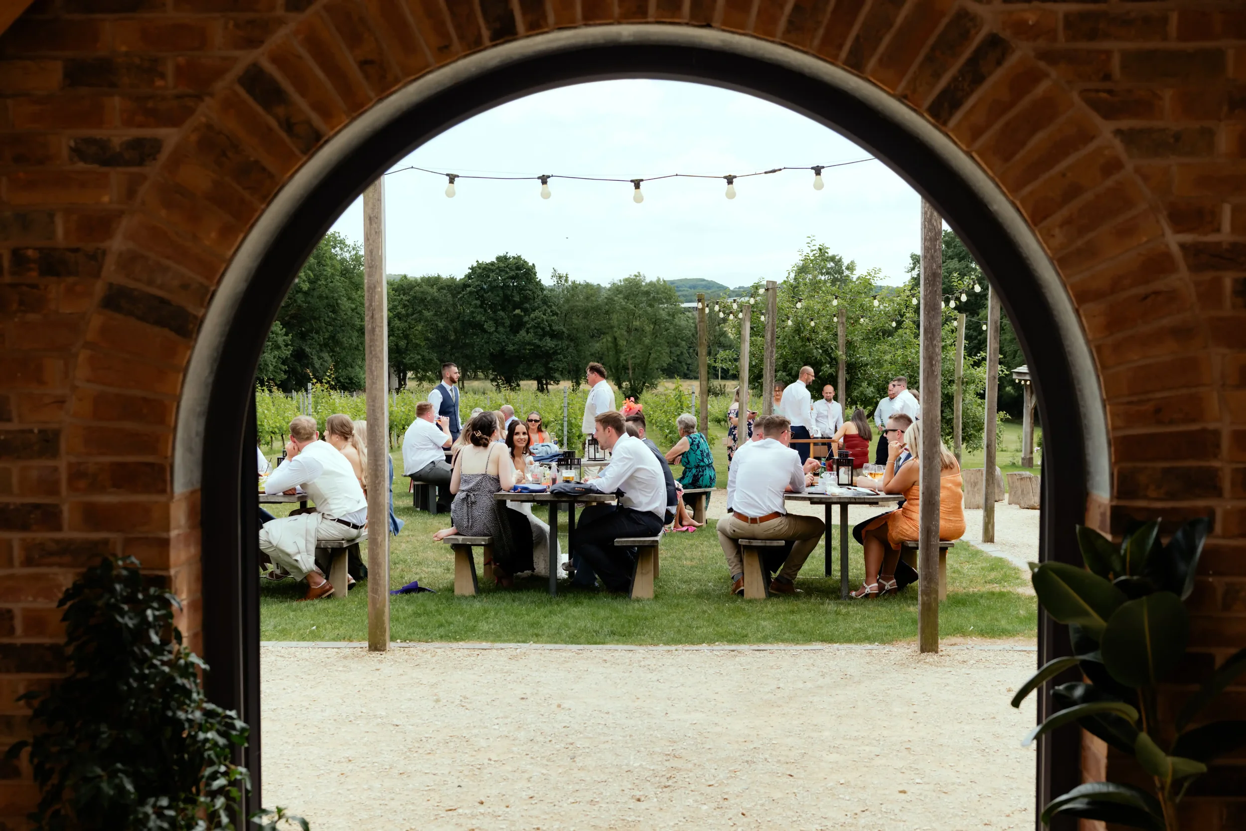 People gathered outdoors at a wedding reception, sitting at tables and mingling on a grassy area, seen through an arched brick doorway.