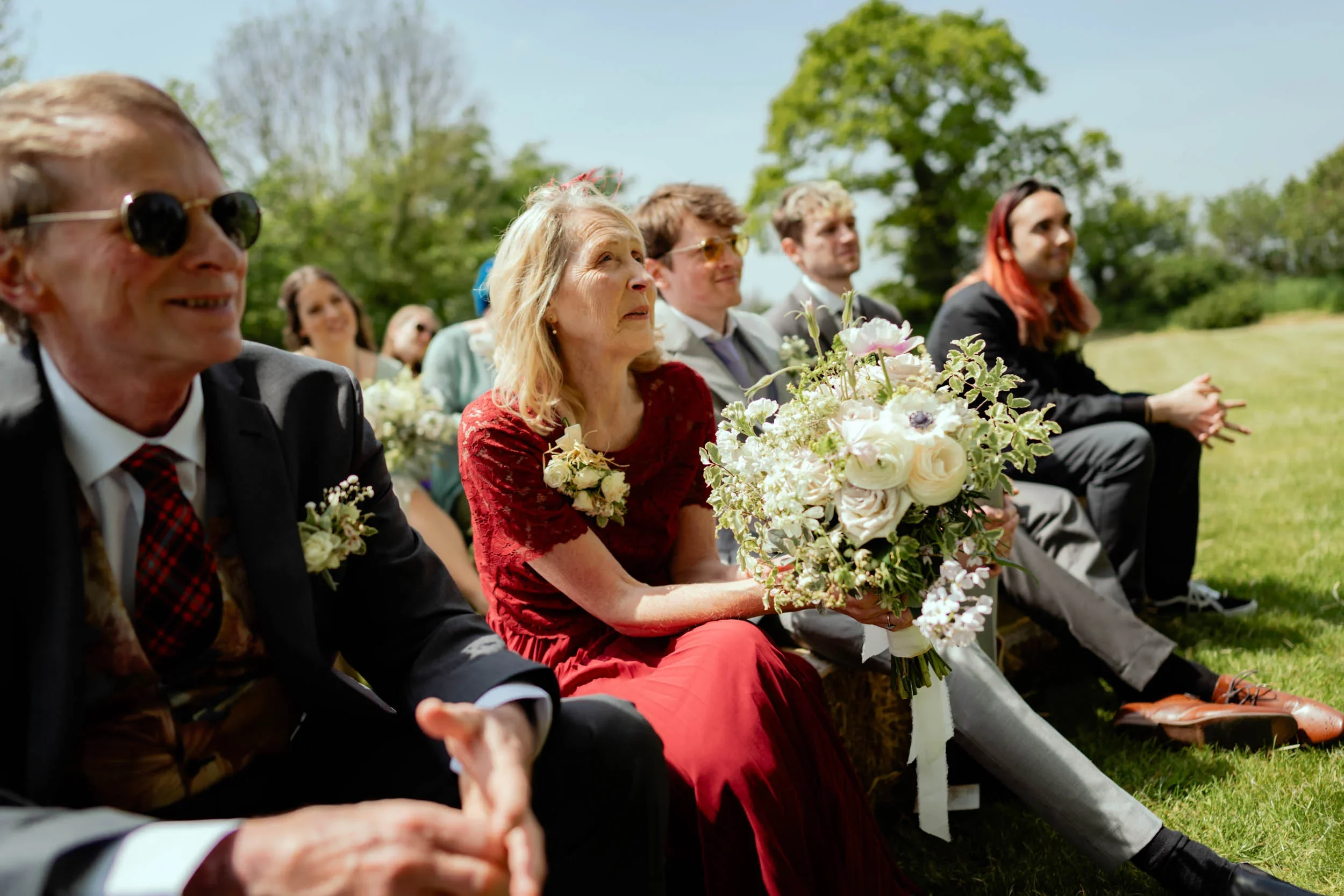 Group of people sitting outdoors at a wedding, dressed in formal attire, with a woman in a red dress holding a large bouquet of white and pink flowers, surrounded by green trees and grass.