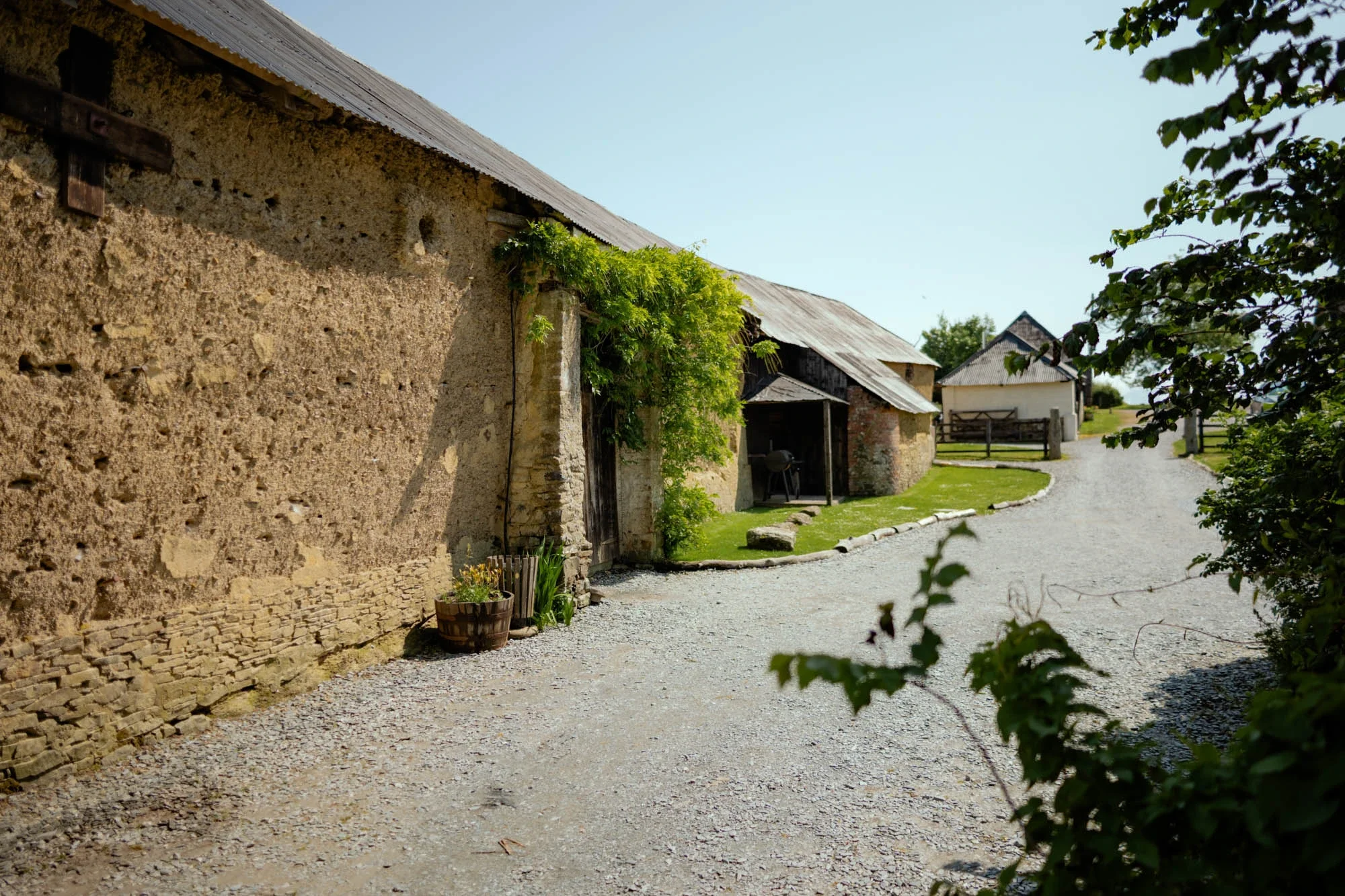 A gravel pathway curves past old stone and wooden farm buildings with metal roofs on a sunny day, greenery surrounds the scene.
