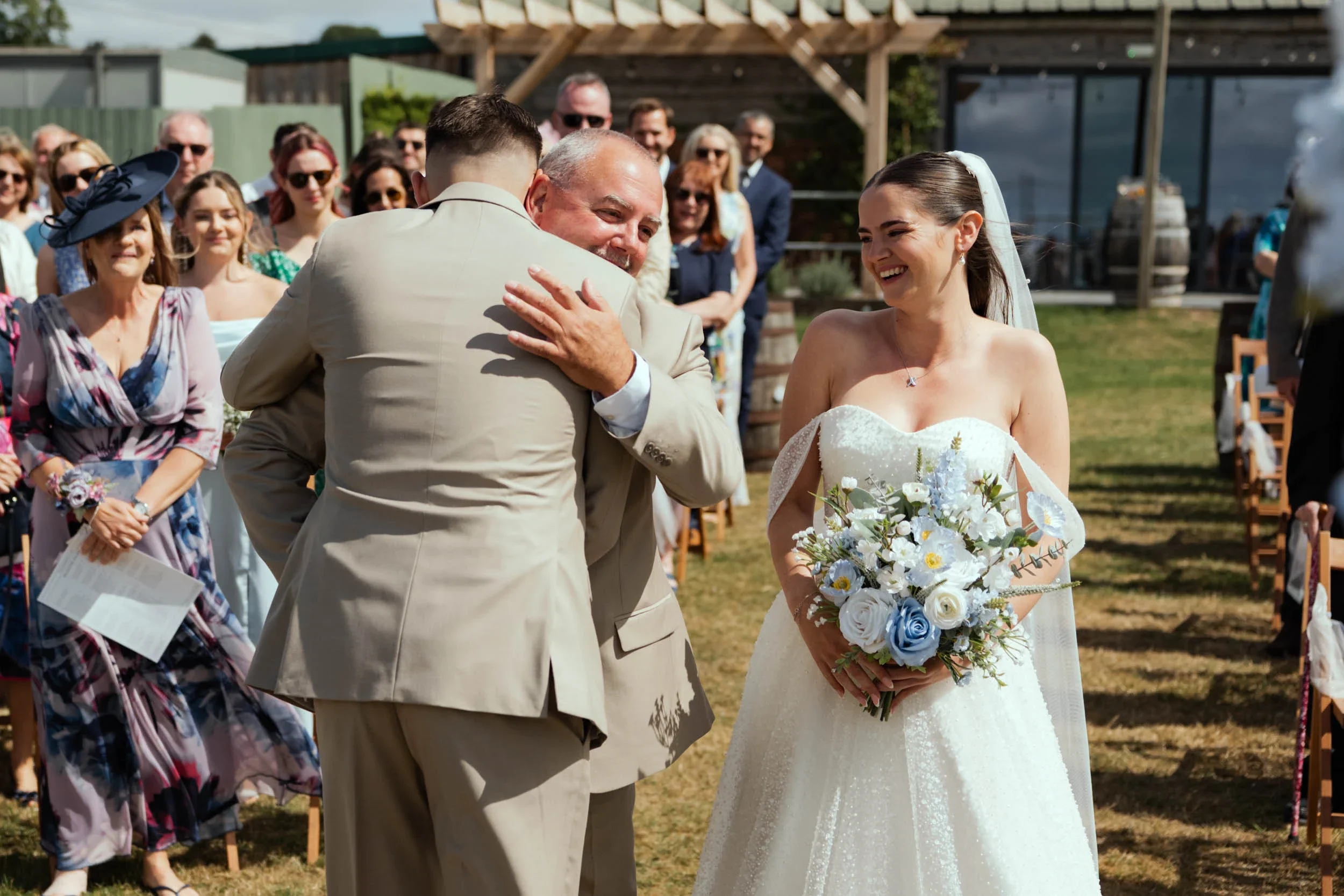 Bride holding a bouquet of white and blue flowers, smiling as she watches a man hug another man at an outdoor wedding ceremony, with guests seated and standing in the background.