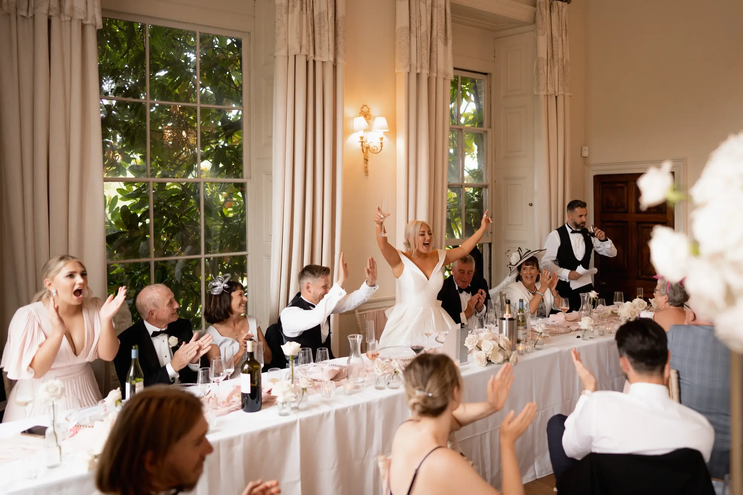 Wedding reception with guests seated at a long table, celebrating and clapping. The bride is standing, smiling, with arms raised, and a man is speaking into a microphone. The room has large windows with curtains, and people are dressed in formal atti