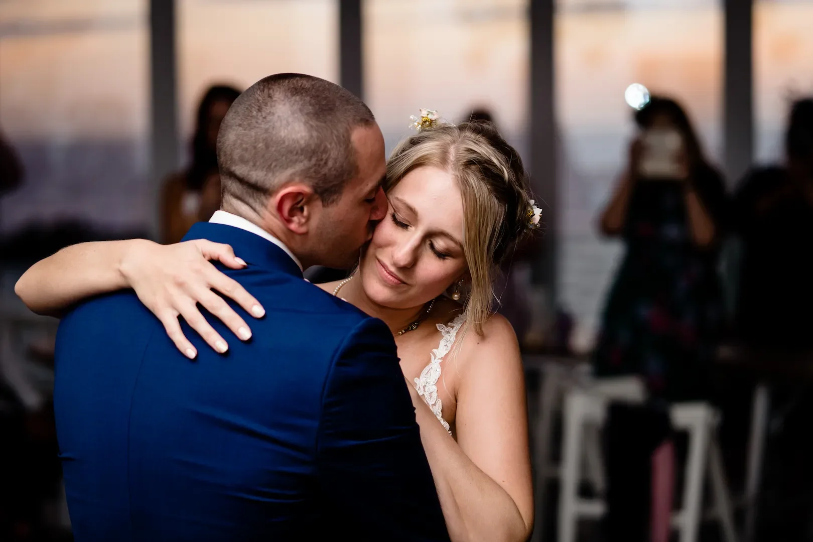 A couple dancing closely at a wedding reception, with the groom wearing a navy suit and the bride wearing a white dress and floral hair accessories.
