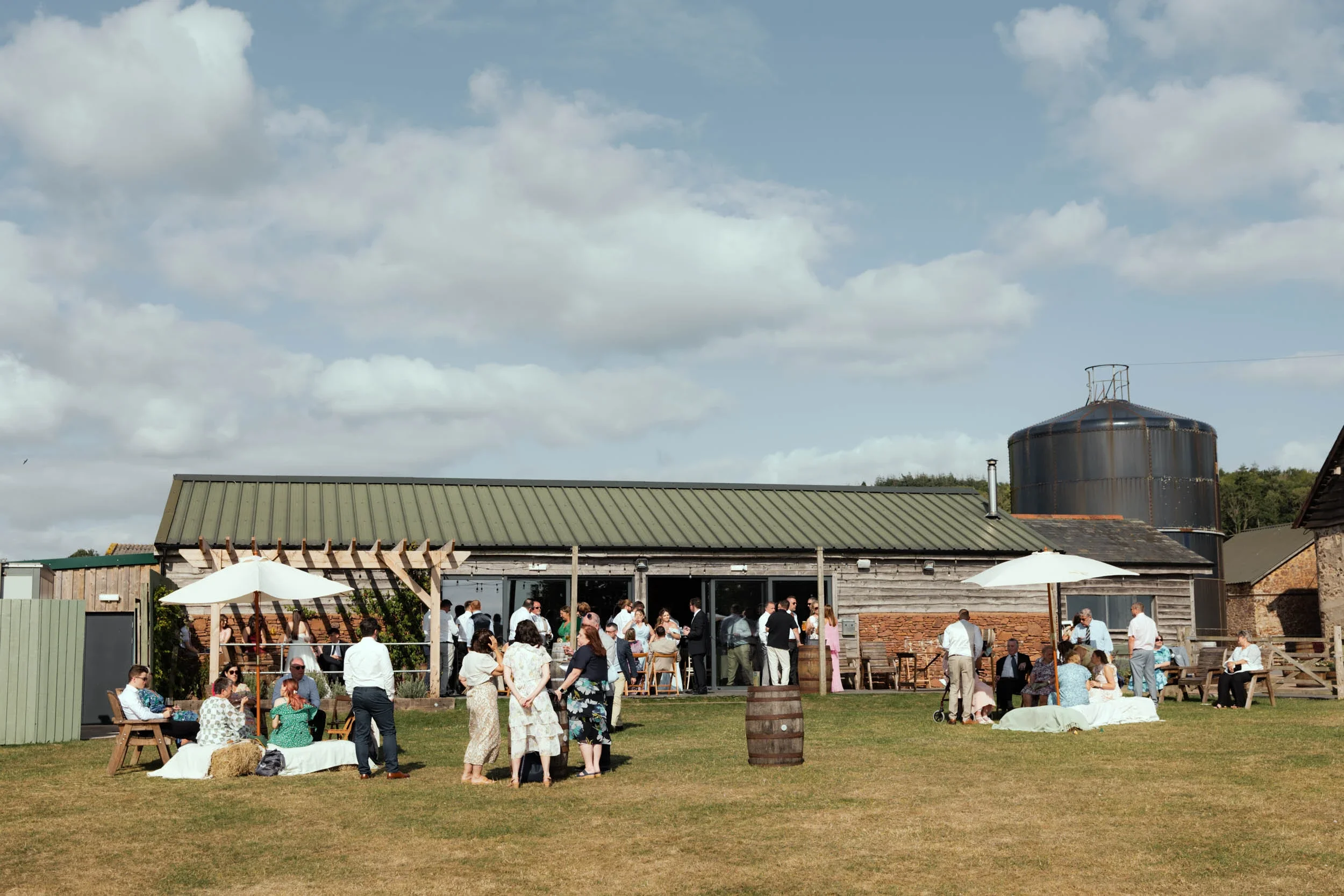 People gathered at an outdoor wedding reception in a rustic farm setting with a barn, large water tank, and umbrellas, enjoying socializing on a sunny day.
