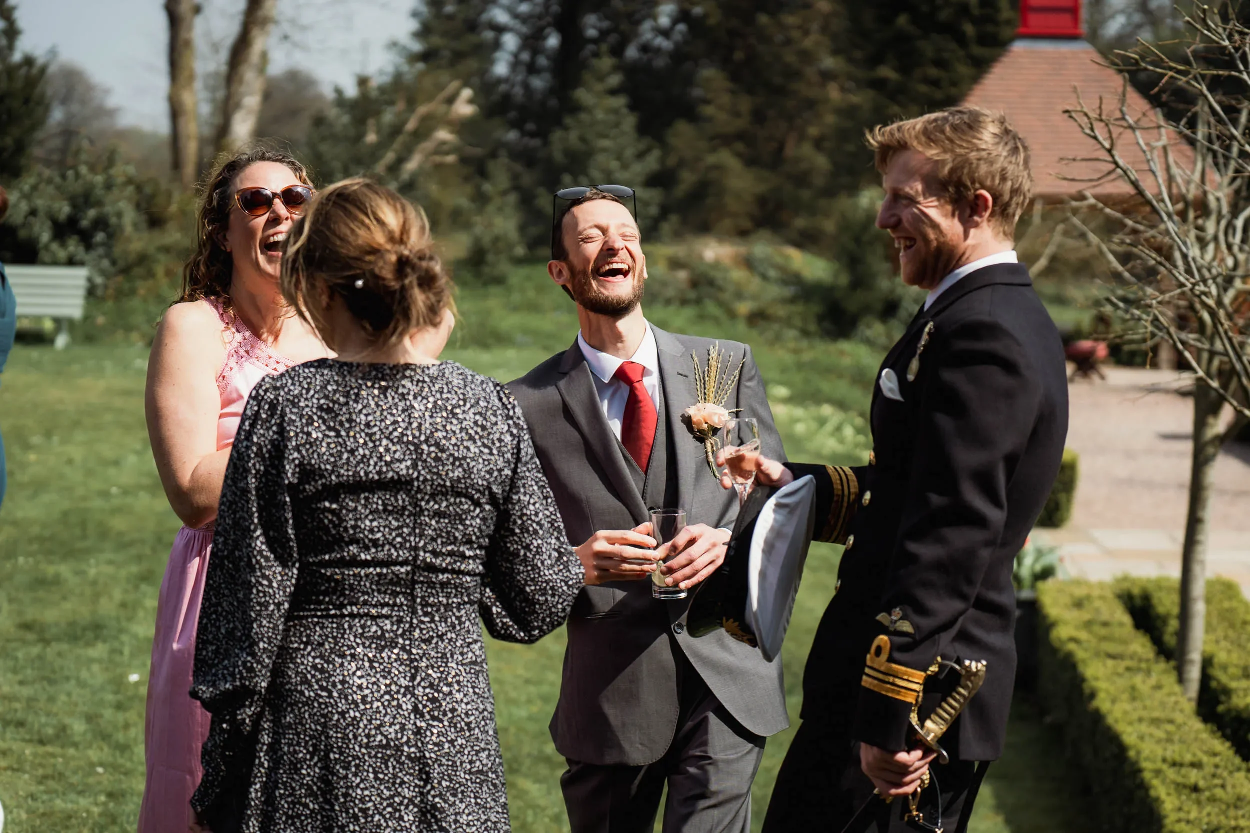 Group of people smiling and laughing outdoors at a social gathering, with a man in a military uniform holding a sword and a woman in a pink dress wearing sunglasses.