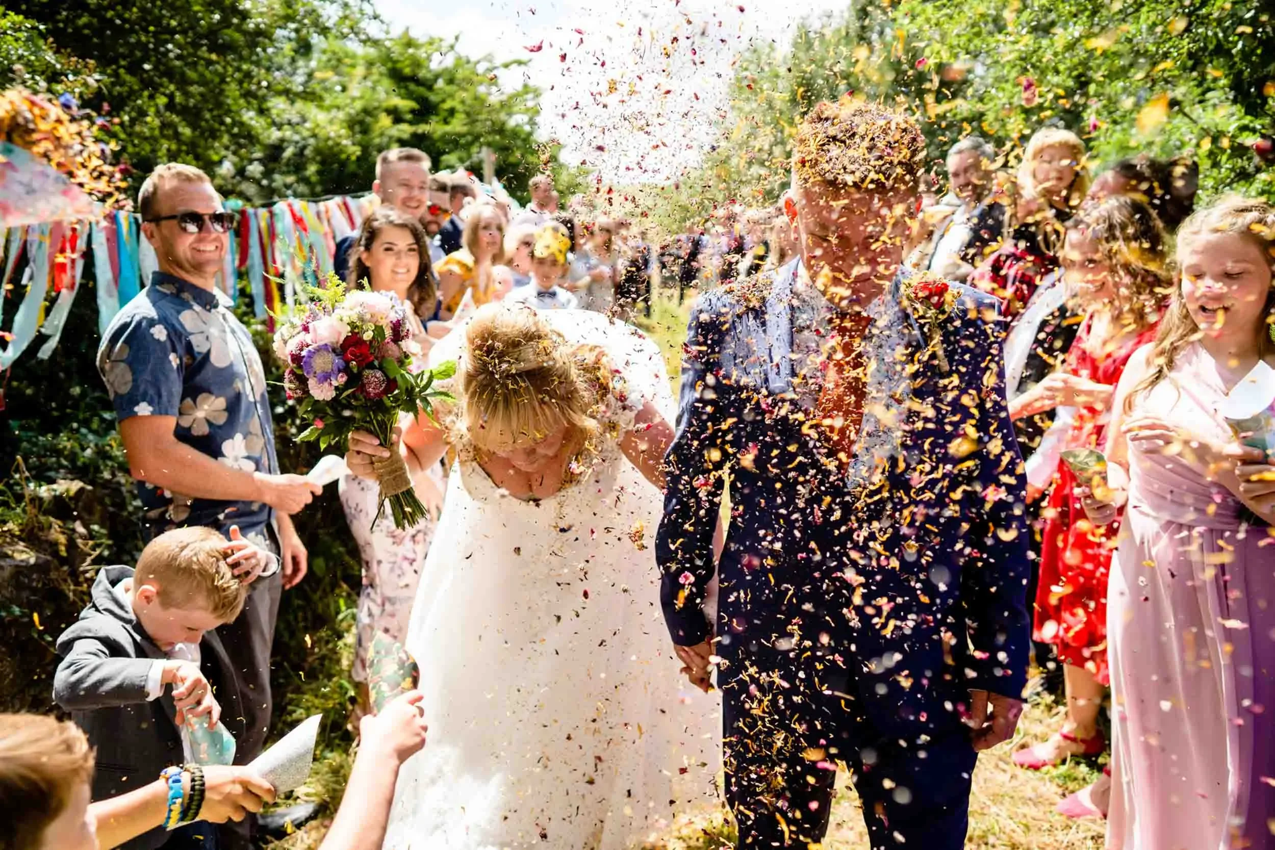 A bride and groom holding hands walking through a crowd after their wedding ceremony as confetti is thrown around them. The bride is holding a bouquet and both are smiling. Guests are cheering and celebrating in the background.