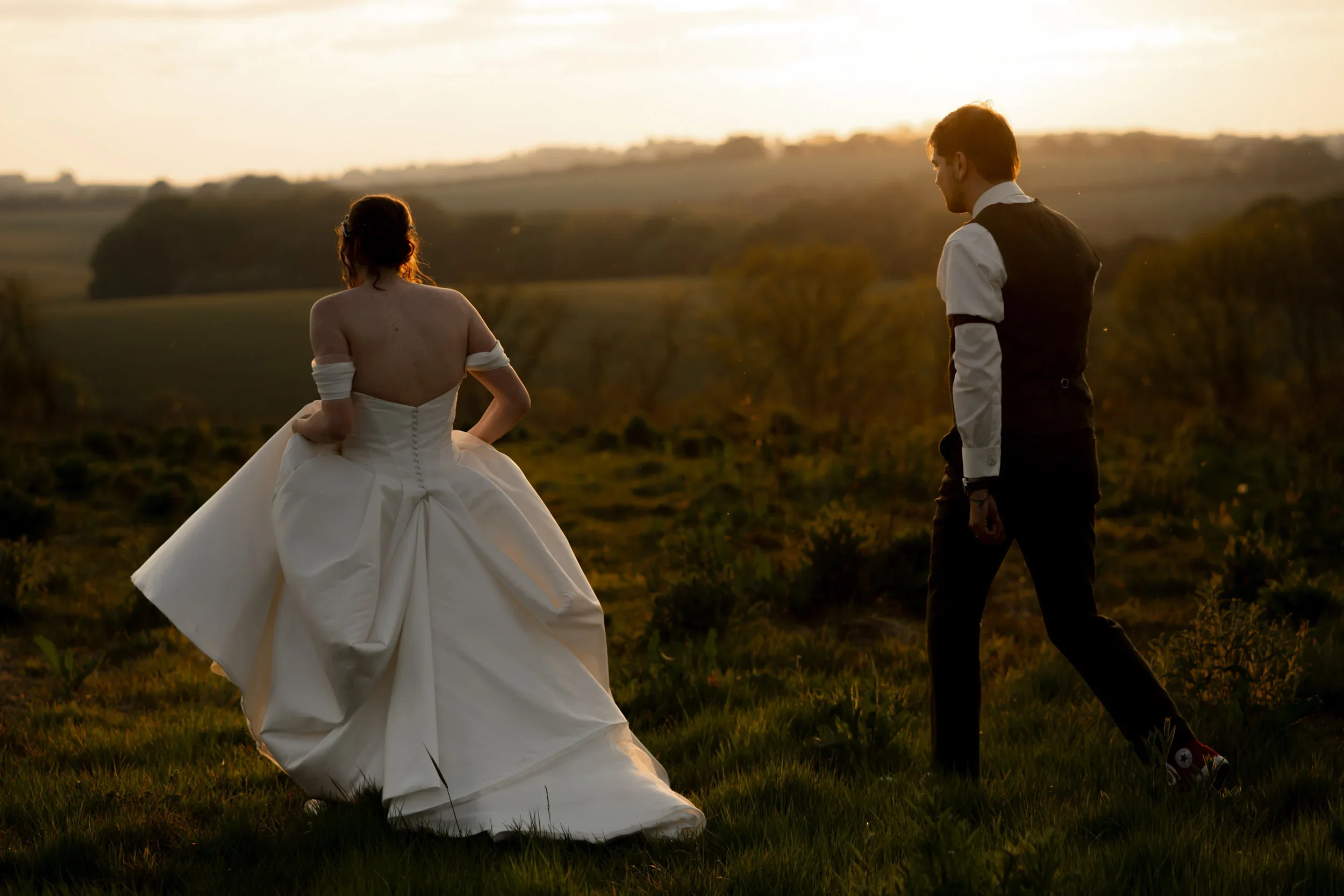 A bride in a white wedding gown and a groom in a black vest and pants standing in a grassy field at sunset.