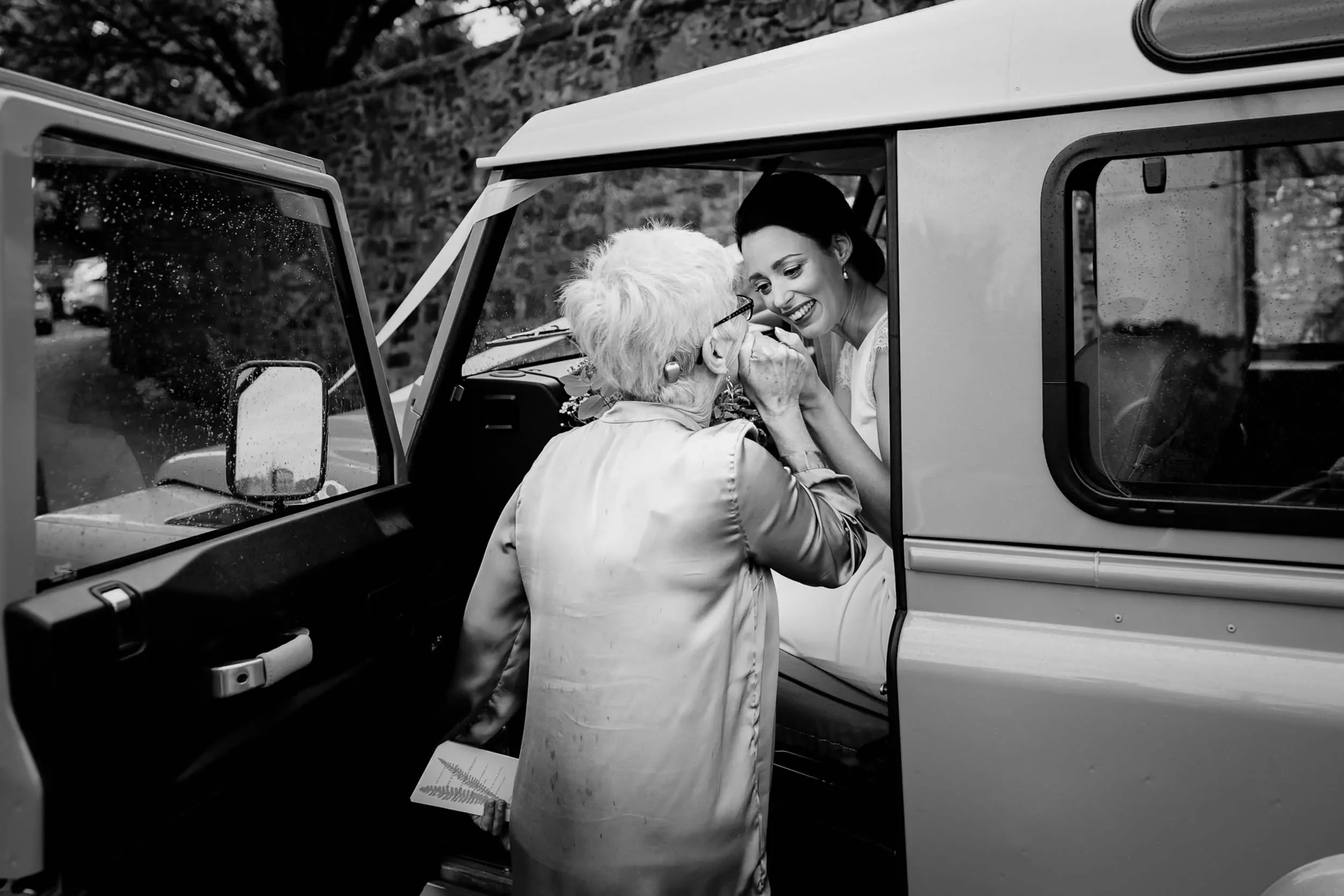 A joyful elderly woman with short white hair and glasses is in a car, holding hands with a smiling young woman with dark hair outside the car window. They are sharing a happy moment, possibly a celebration like a wedding or special occasion, on a rai