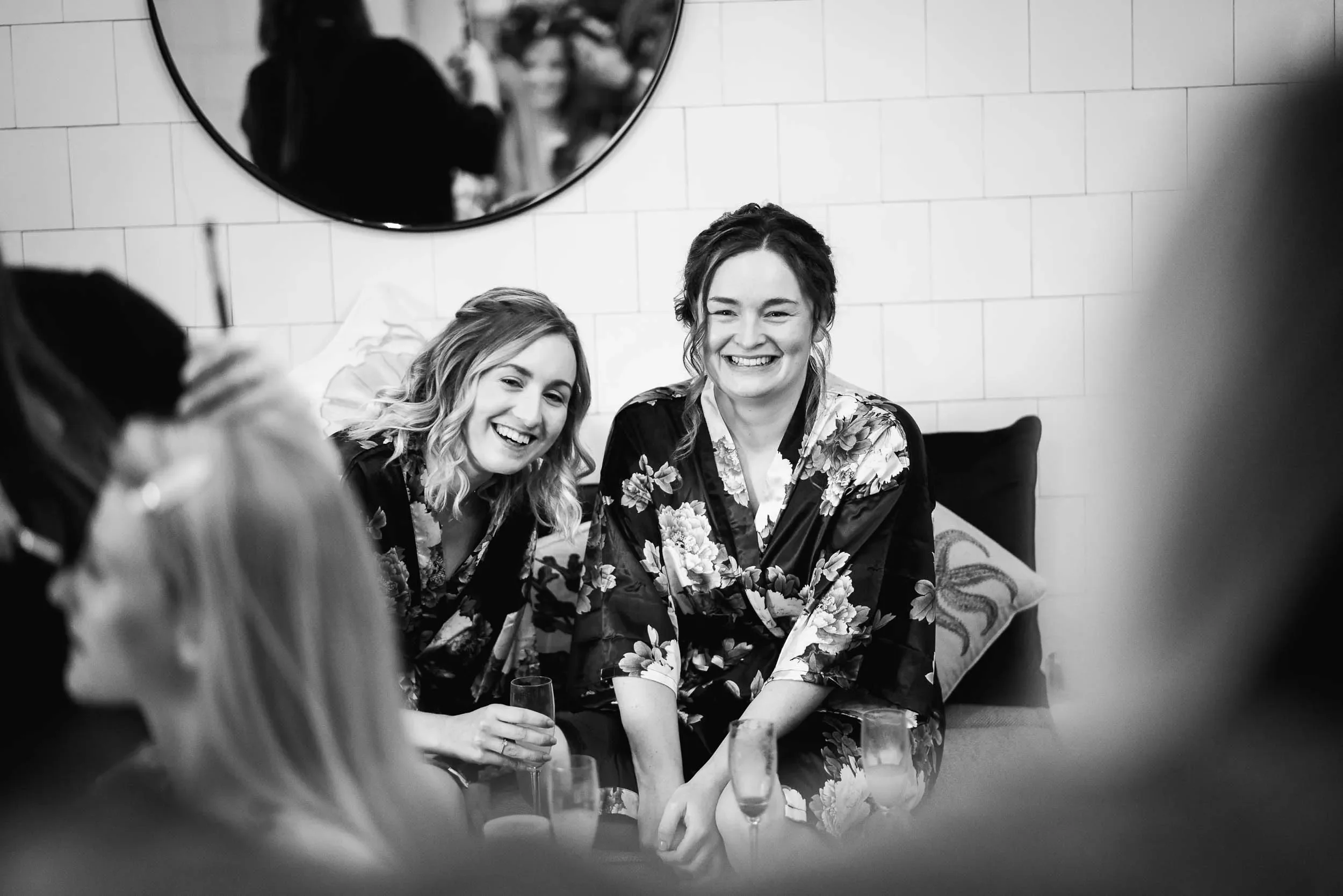 Two women in floral robes laughing and holding champagne glasses at a social gathering, seen in a black-and-white photo.