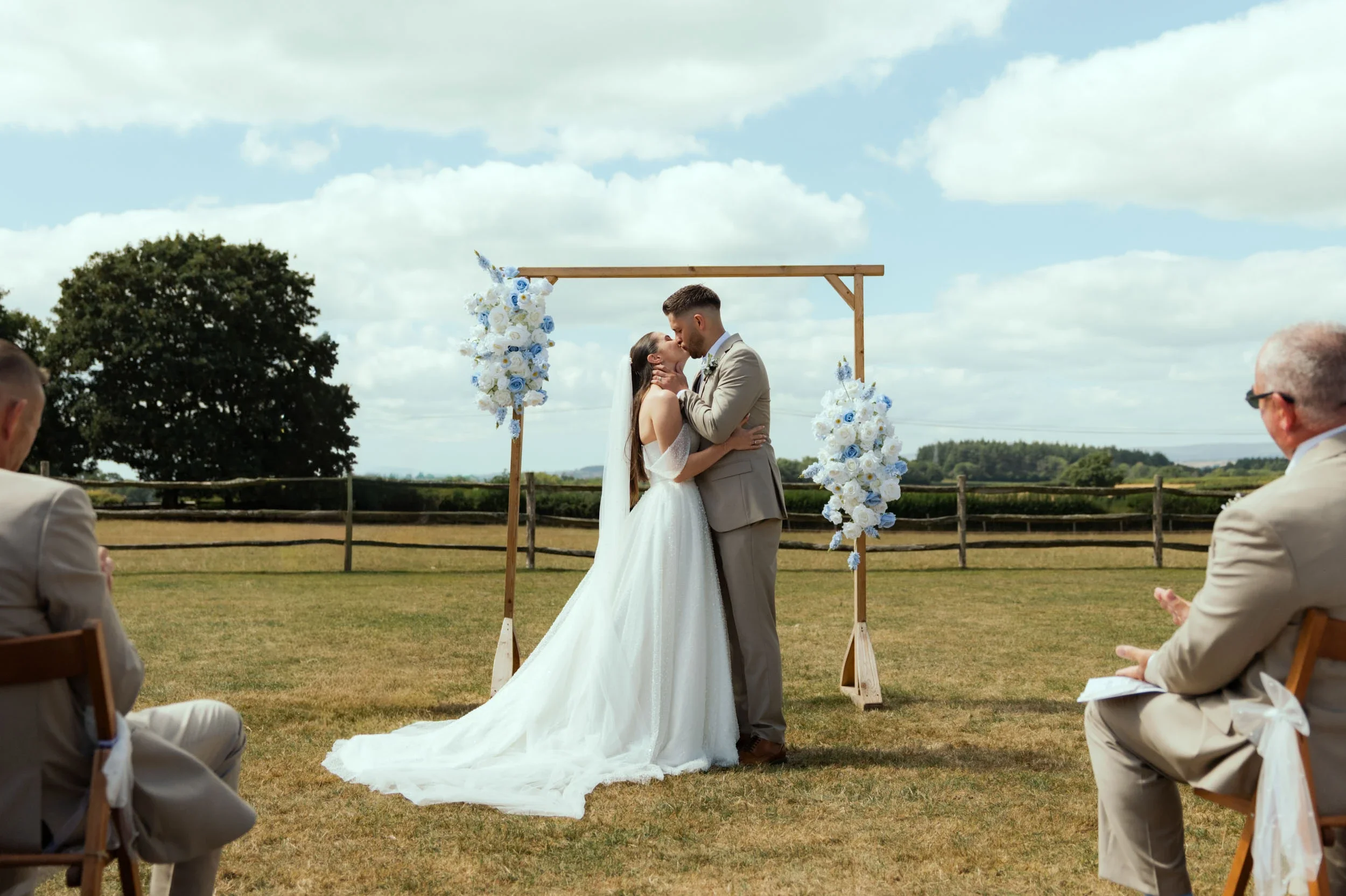 A bride and groom kissing during an outdoor wedding ceremony on a grassy field with a wooden arch decorated with white and blue flowers, and guests seated on wooden chairs watching.