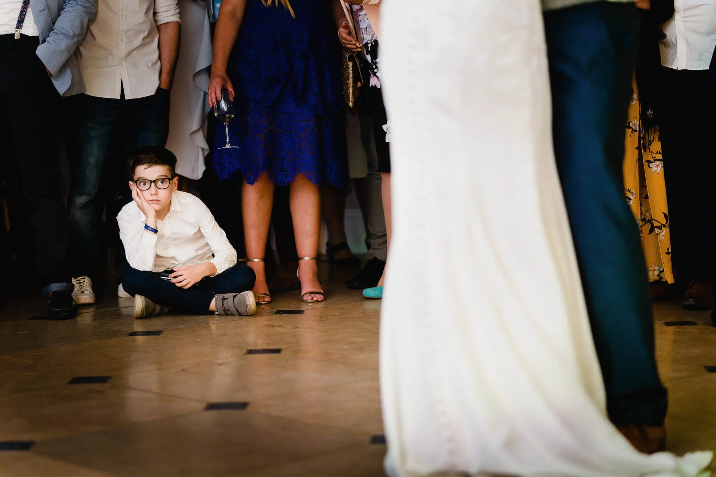 Young boy sitting on the floor at a social event, resting his head on his hand, surrounded by standing adults dressed in formal attire.