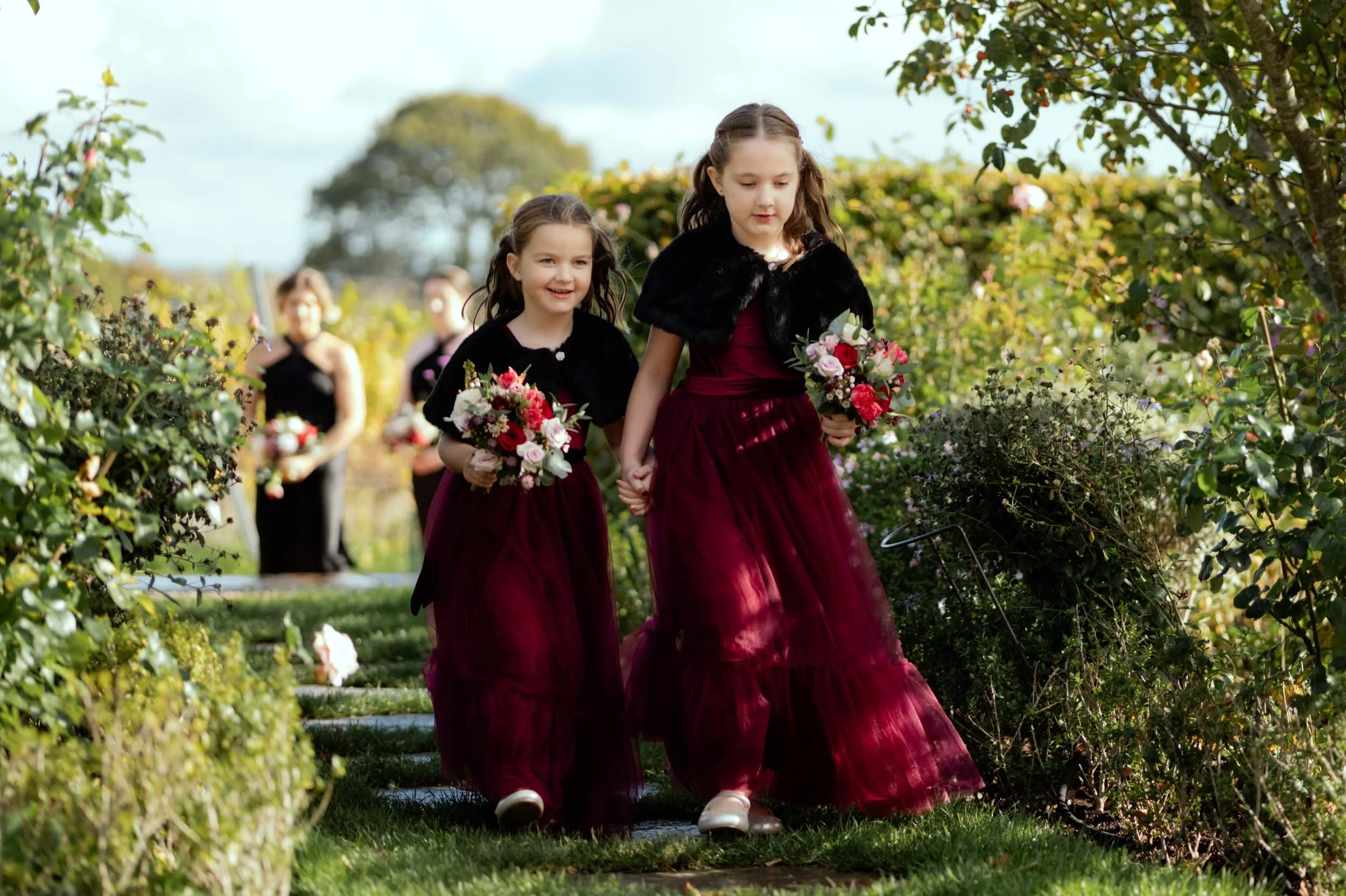 Group of young girls in matching maroon dresses and black shawls walking through a garden, holding bouquets of flowers, with trees in the background.