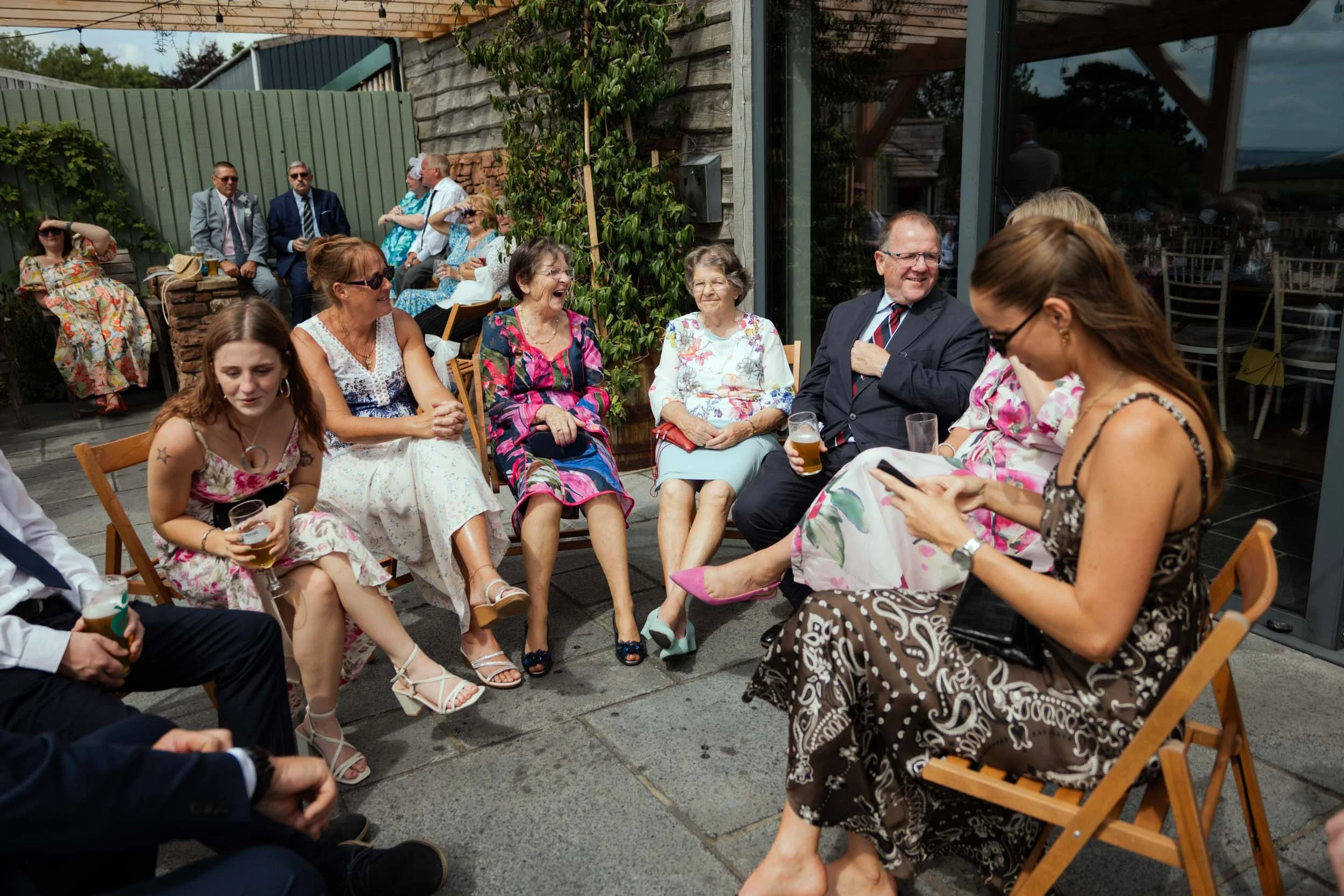 Group of people enjoying a social gathering, sitting on chairs outside on a patio, some holding drinks, laughing, talking, and looking at a woman who is using her phone.