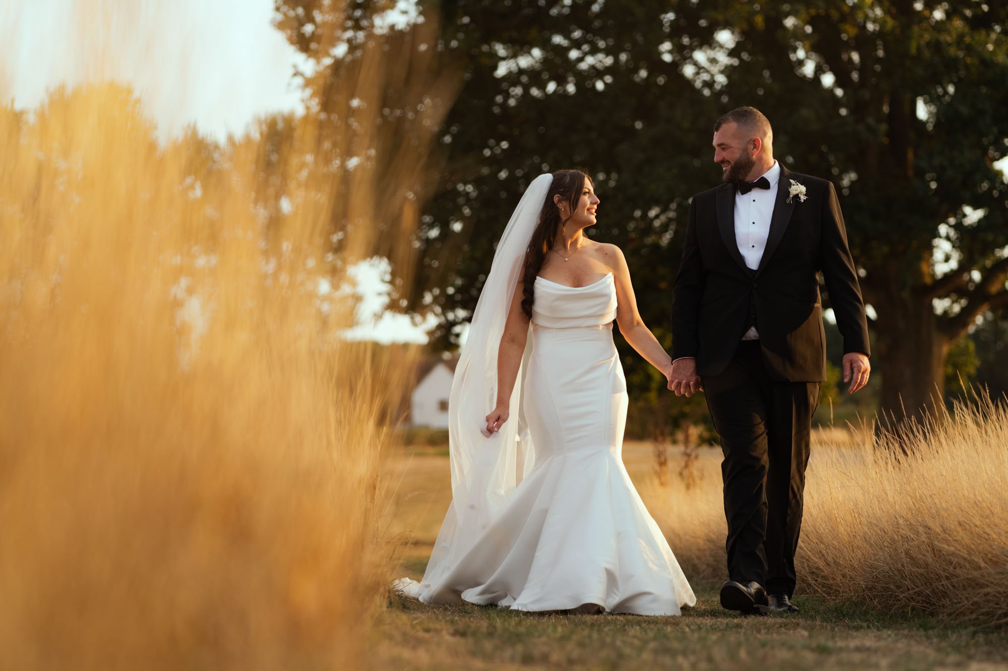 A bride and groom walking hand in hand outdoors during sunset, with dry tall grass on either side and trees in the background.