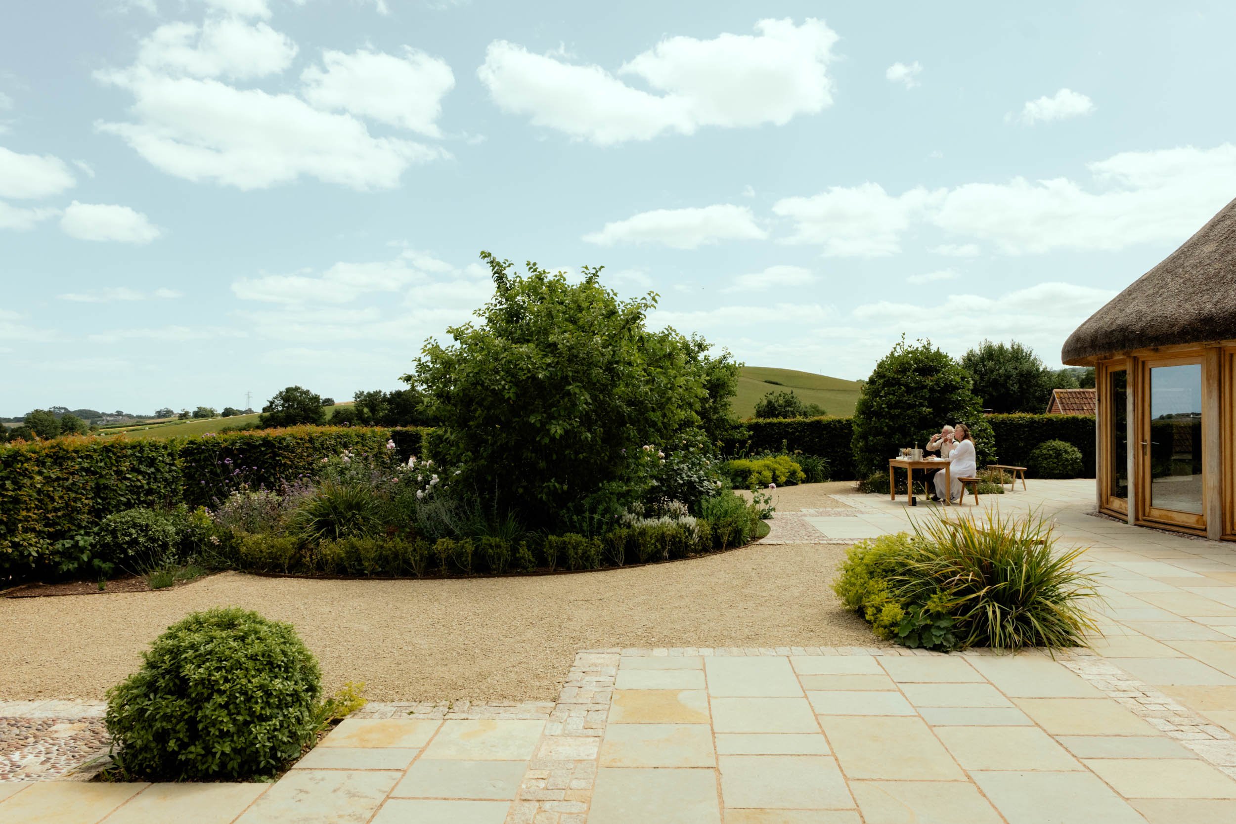 An outdoor patio with a stone floor, surrounded by greenery and shrubs. There is a large tree and a hedge, with rolling hills and a blue sky with clouds in the background. Two people are seated at a table on the patio, enjoying a meal or drinks.