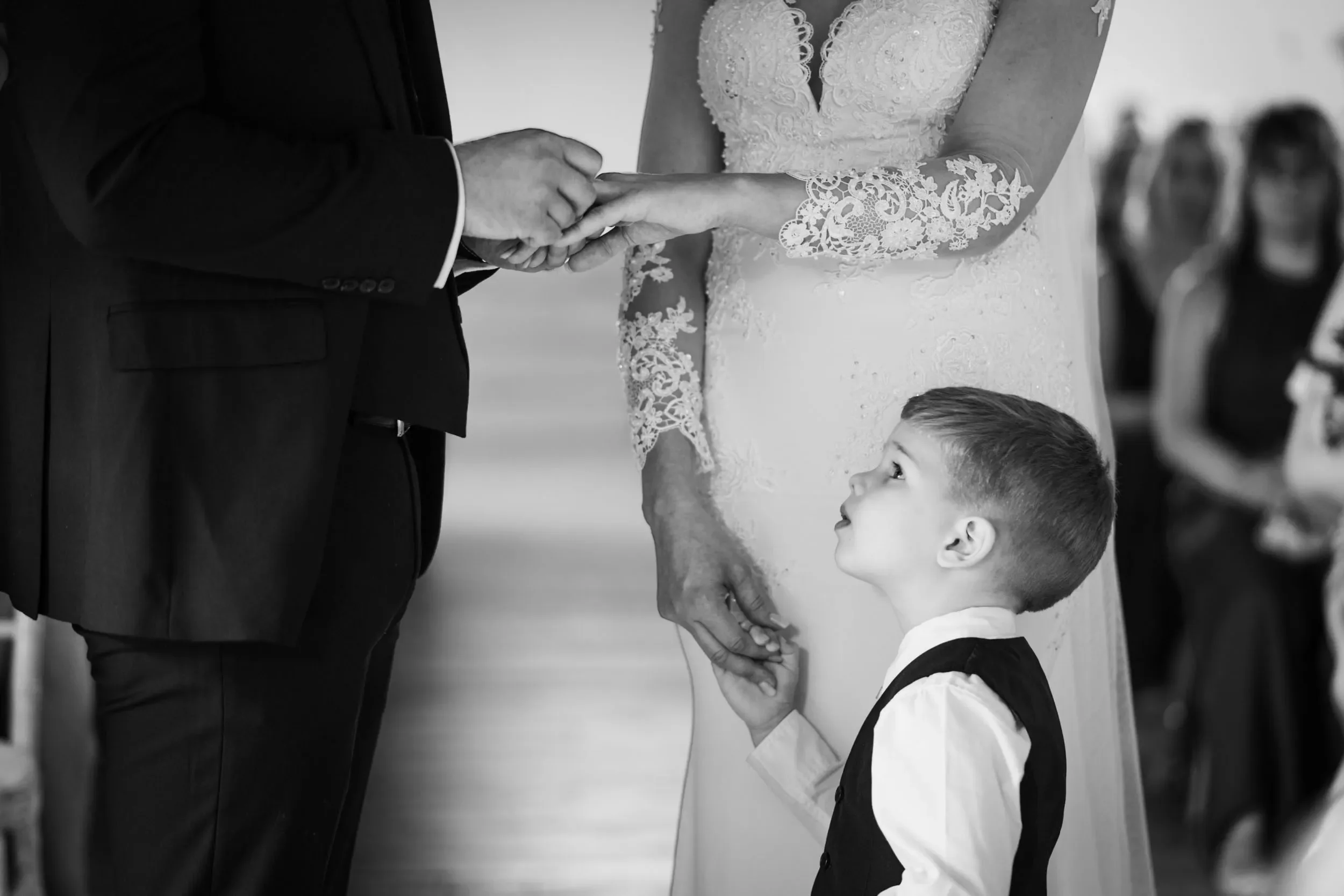A young boy looks up at a couple exchanging wedding vows while holding the woman's hand, with the man in a dark suit and the woman in a lace wedding dress, during a wedding ceremony.