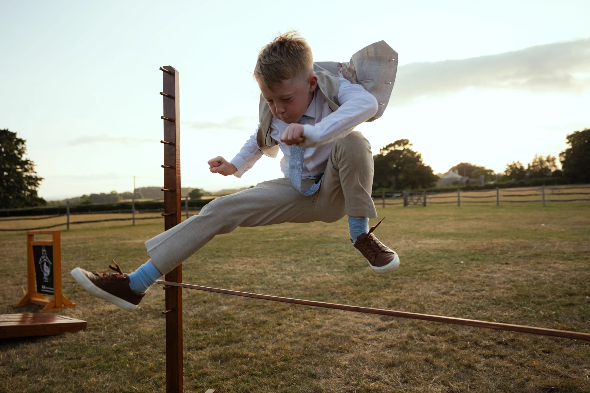 Young boy in formal attire jumping over a wooden obstacle in a field at sunset.