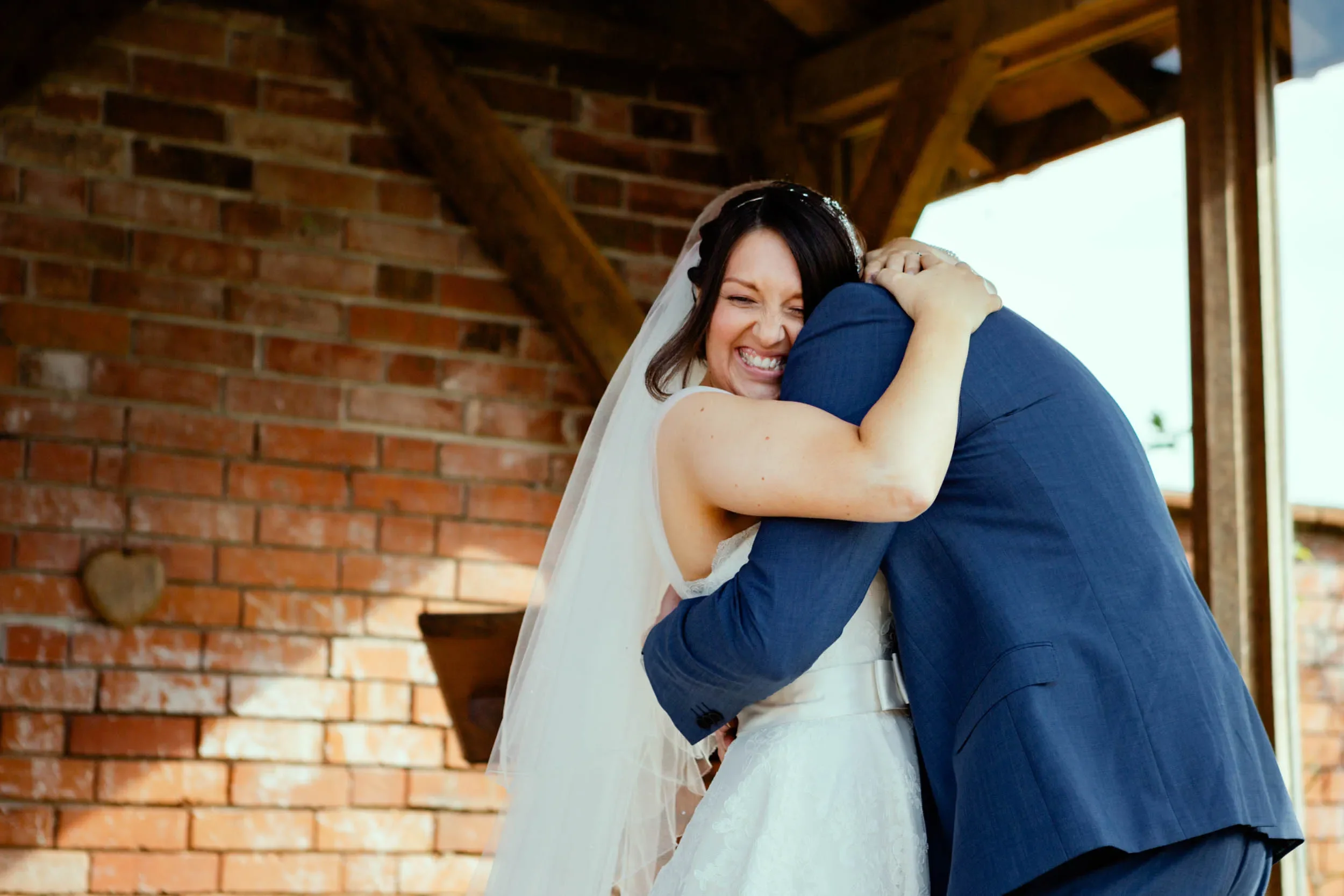A bride smiling happily and hugging a man in a navy suit, with a brick wall and wooden beams in the background.
