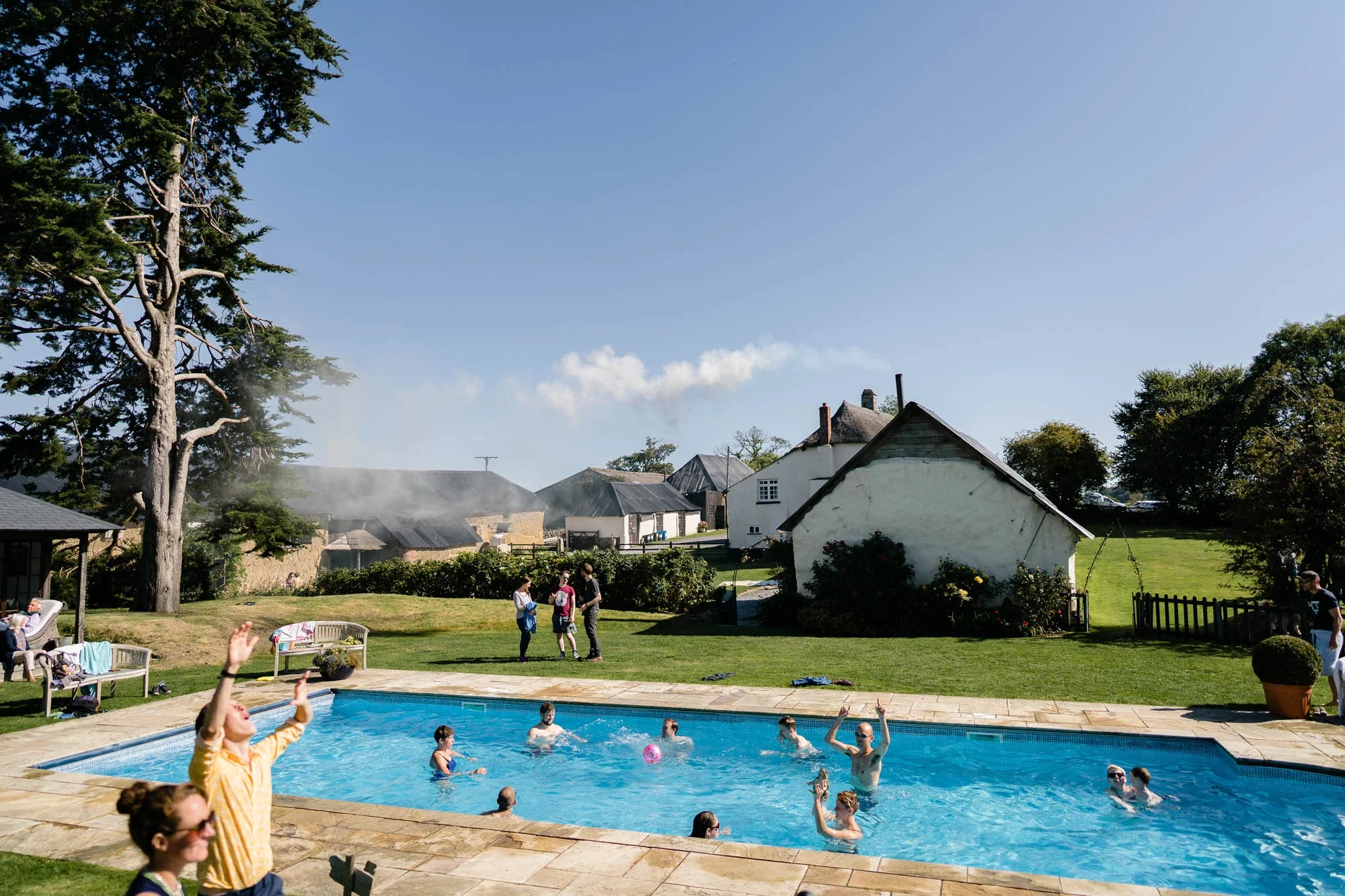 A group of people playing volleyball in a swimming pool in a backyard with green grass, trees, and houses in the background on a sunny day.