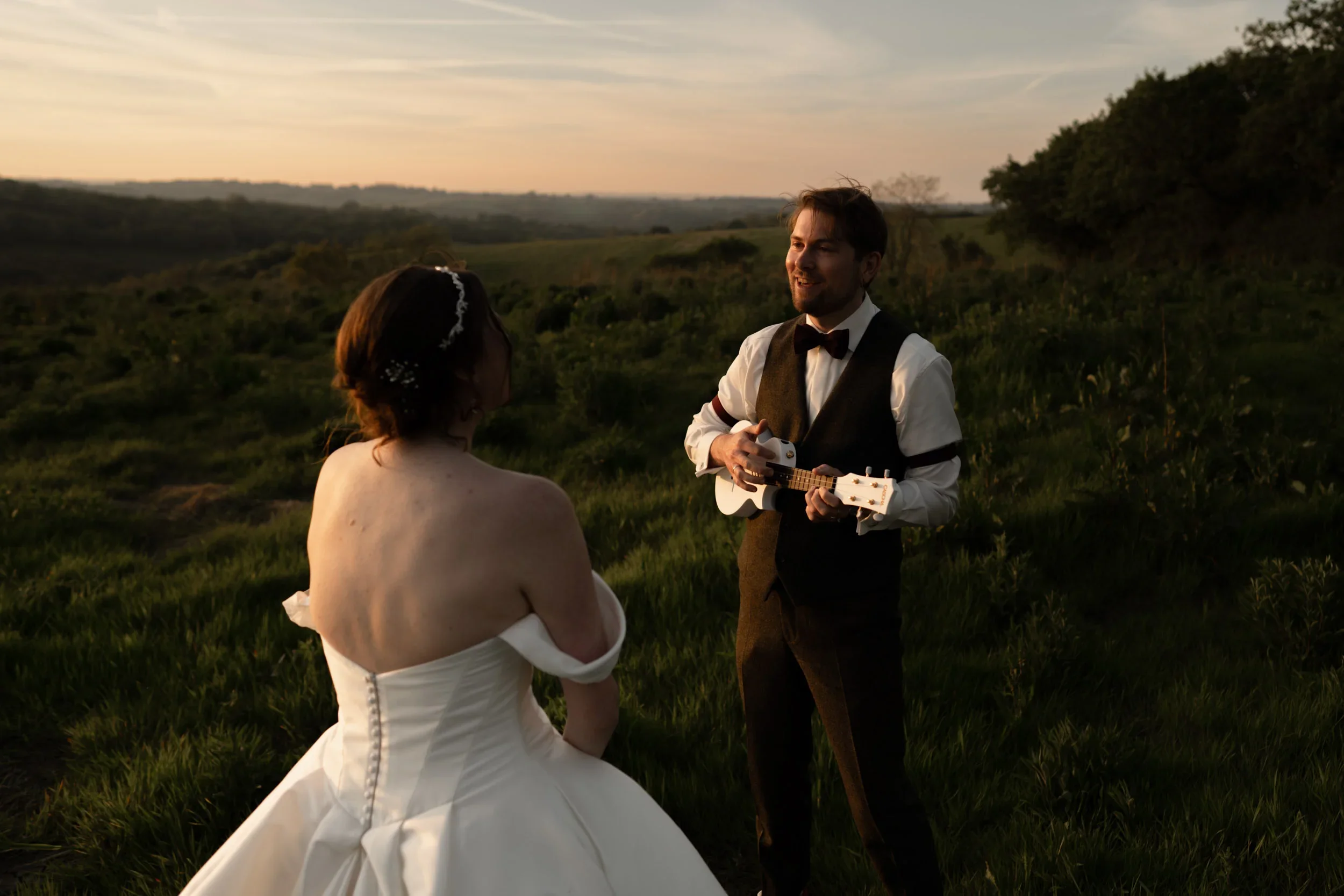 A man in a tuxedo playing a ukulele serenades a woman in a wedding dress outdoors during sunset.