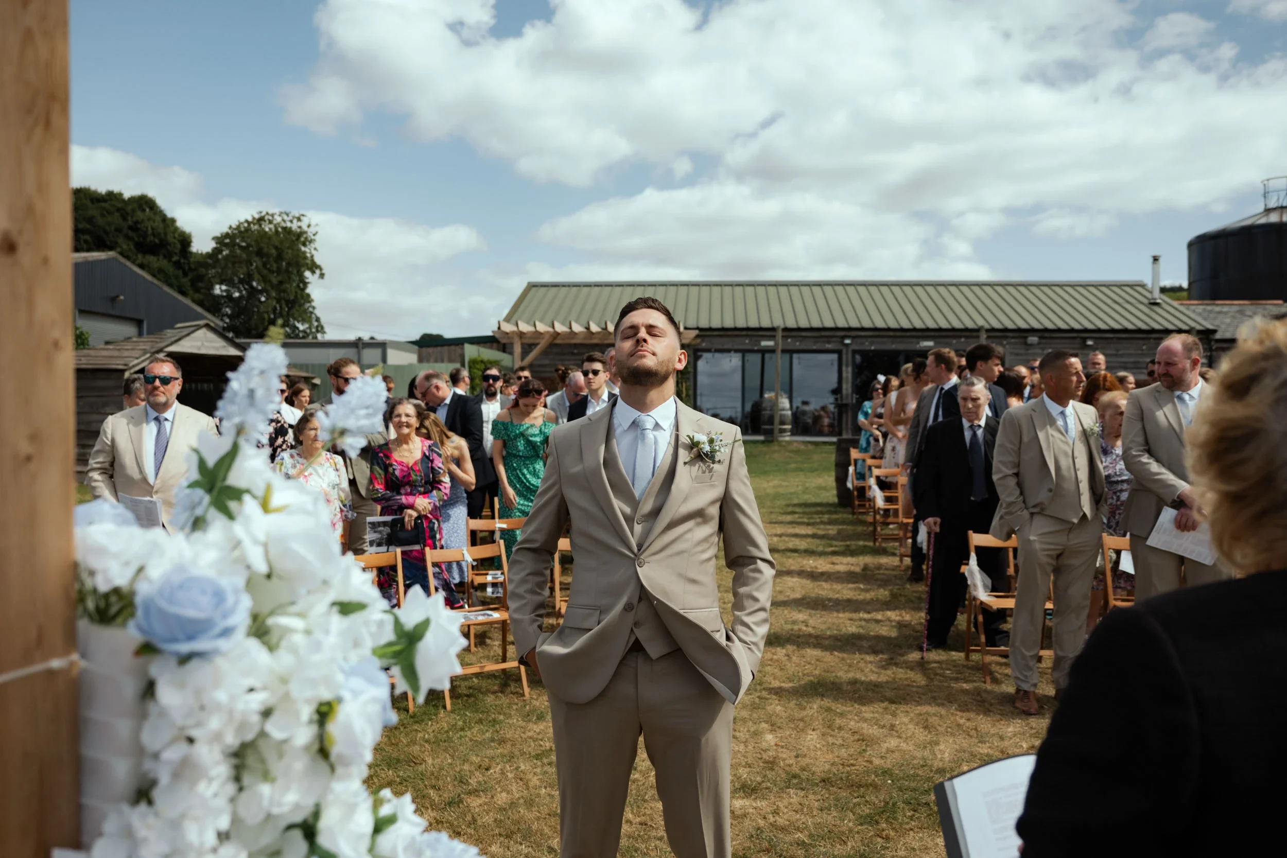 Groom in beige suit with a boutonniere, standing with hands in pockets at outdoor wedding ceremony, surrounded by seated guests on grassy field under cloudy sky.