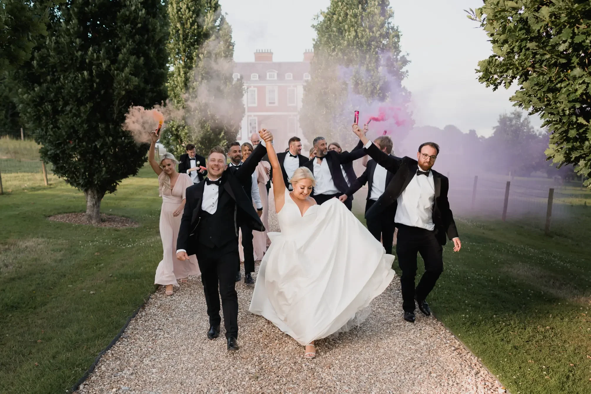 A group of wedding guests, including a bride and groom, celebrating outdoors on a gravel path with trees and a large house in the background. The bride is being lifted by the groom, and the party is holding pink smoke bombs.