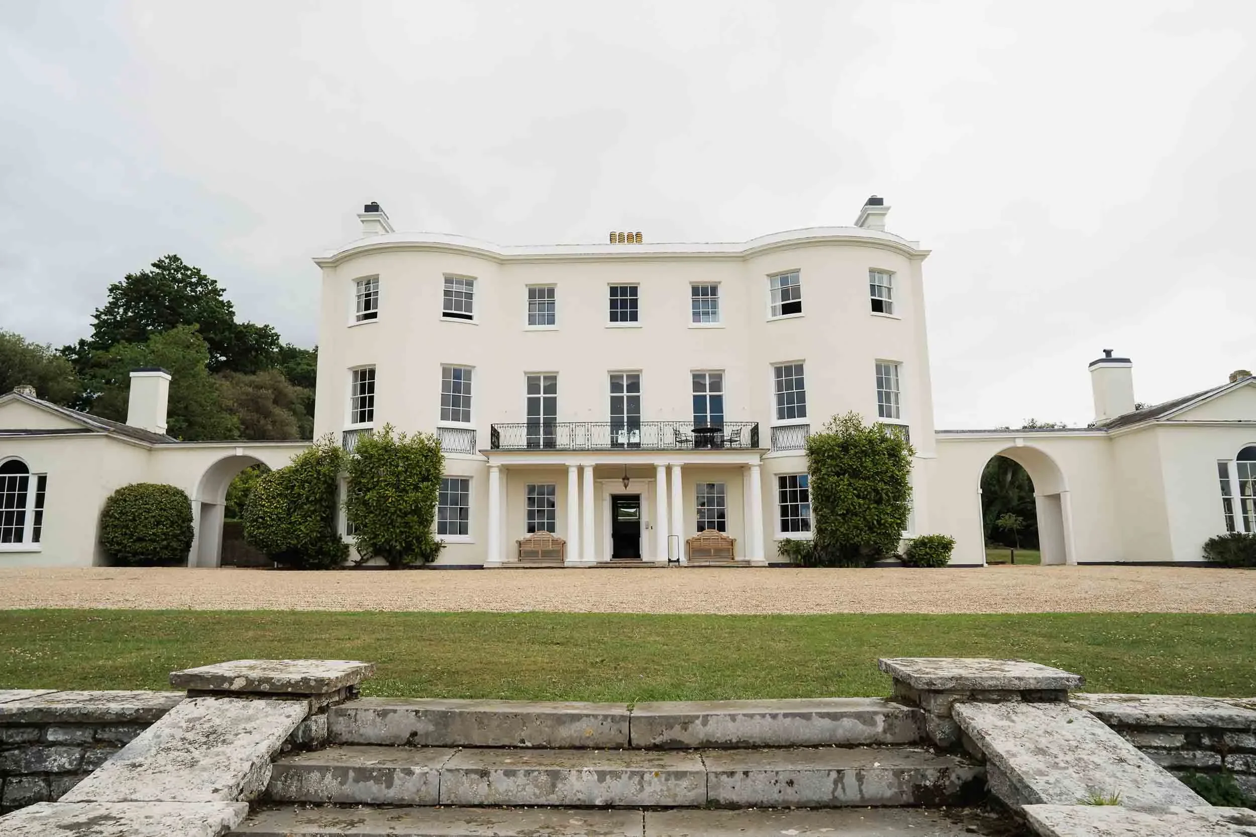 A large white mansion with multiple windows, columns, and a balcony, surrounded by greenery and stone steps in the foreground.