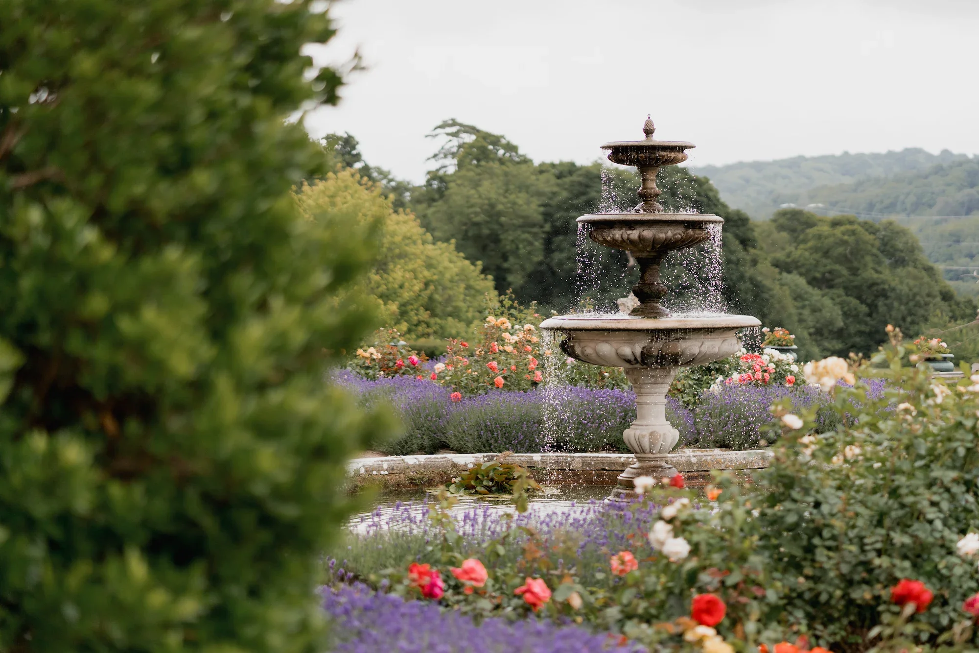 Stone fountain in a garden surrounded by colorful flowers and green trees with hills in the background.