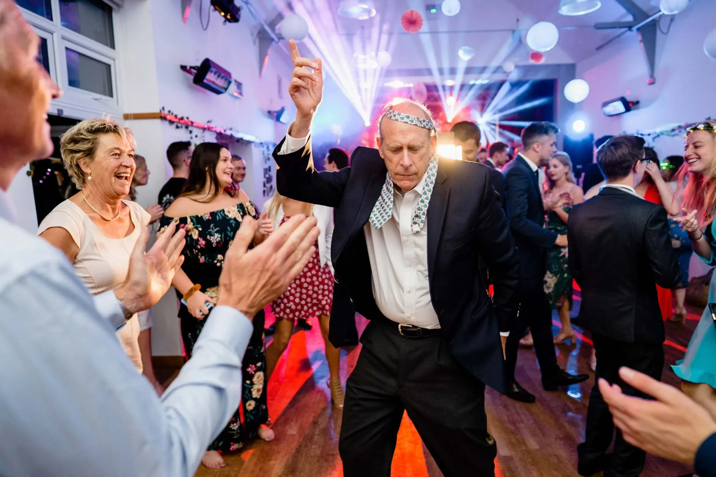 Older man dancing with a headband at a lively party, surrounded by smiling and clapping guests in colorful attire, with dynamic lighting and decorations in the background.