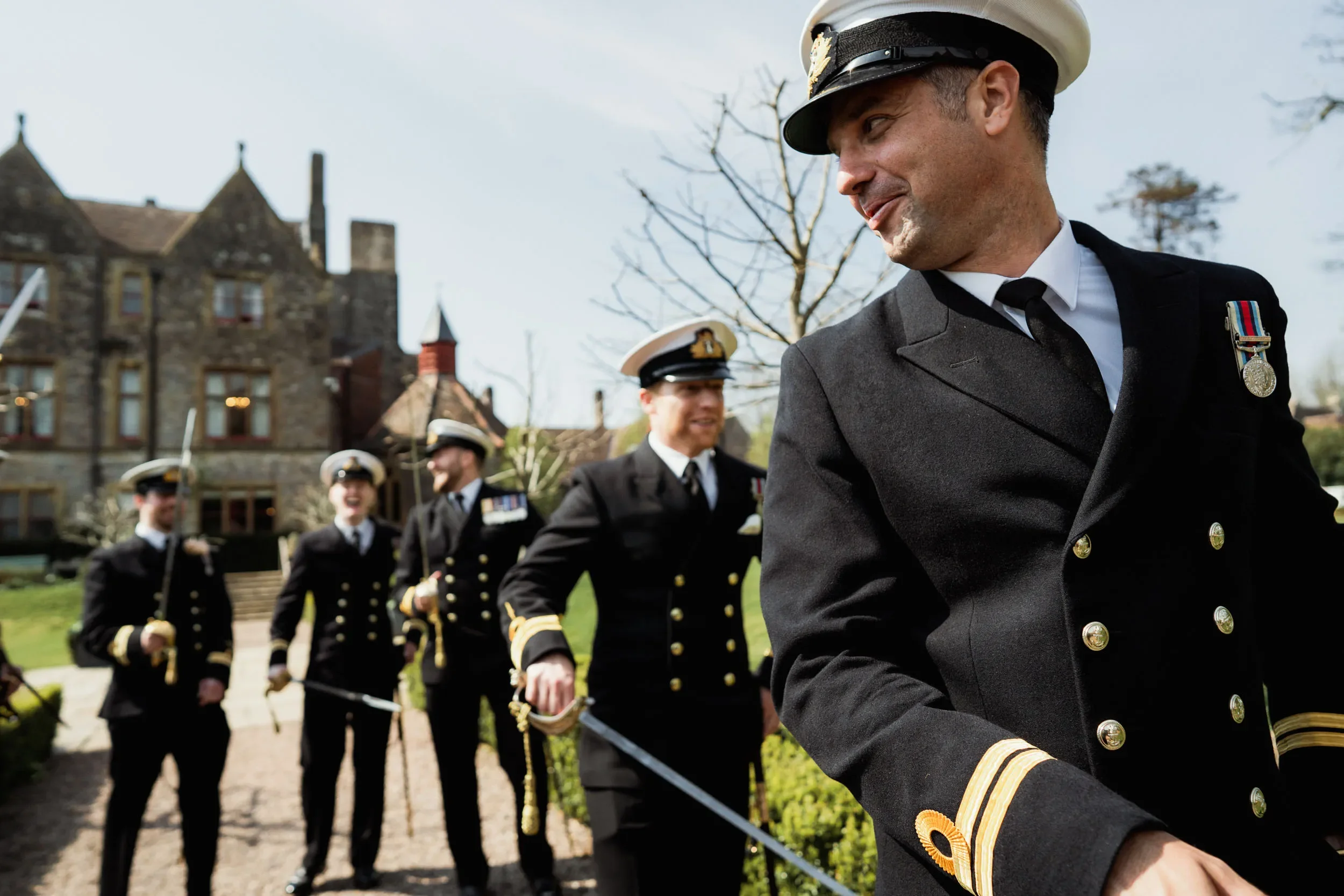 A group of men in navy uniforms with medals, walking outdoors with smiles, on a sunny day, in front of a large, old stone house and trees.