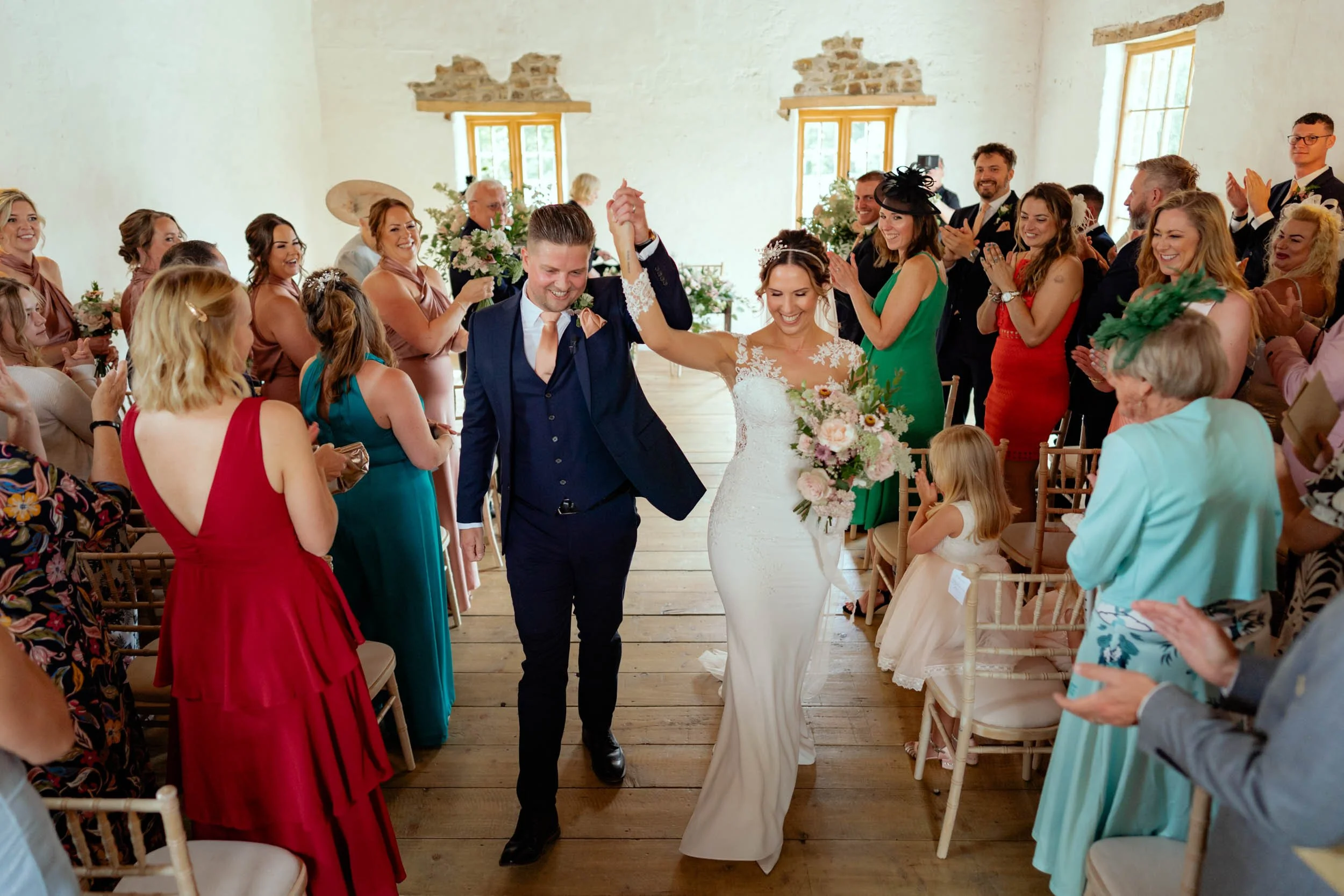 A newlywed couple walking down the aisle hand in hand, smiling. The bride is in a white wedding dress holding a bouquet, and the groom is in a navy suit. Guests are standing, clapping, and smiling in a rustic indoor wedding setting.