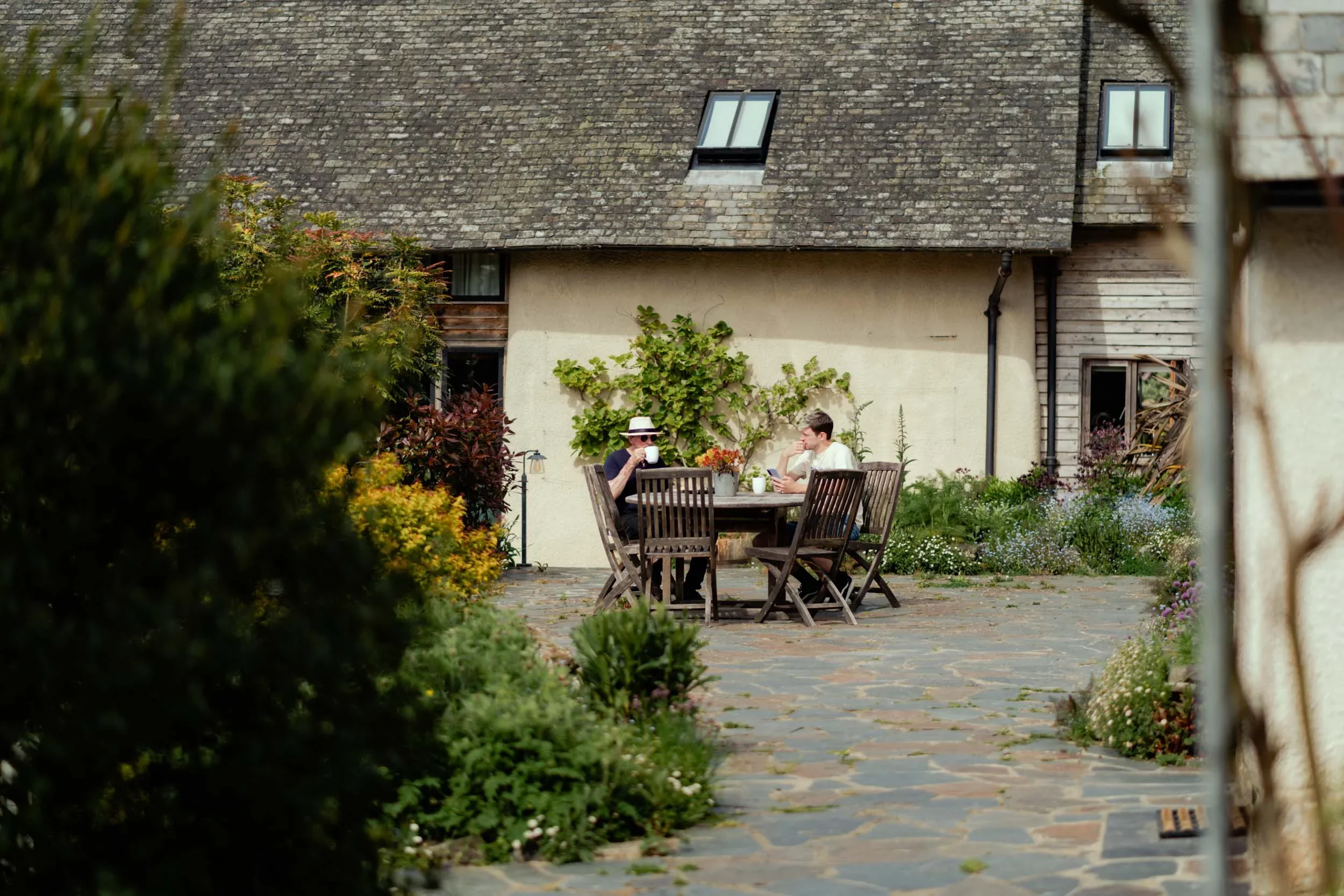 Two people sitting at a round wooden table in a garden, one drinking coffee and the other using a tablet, with a house and plants in the background.