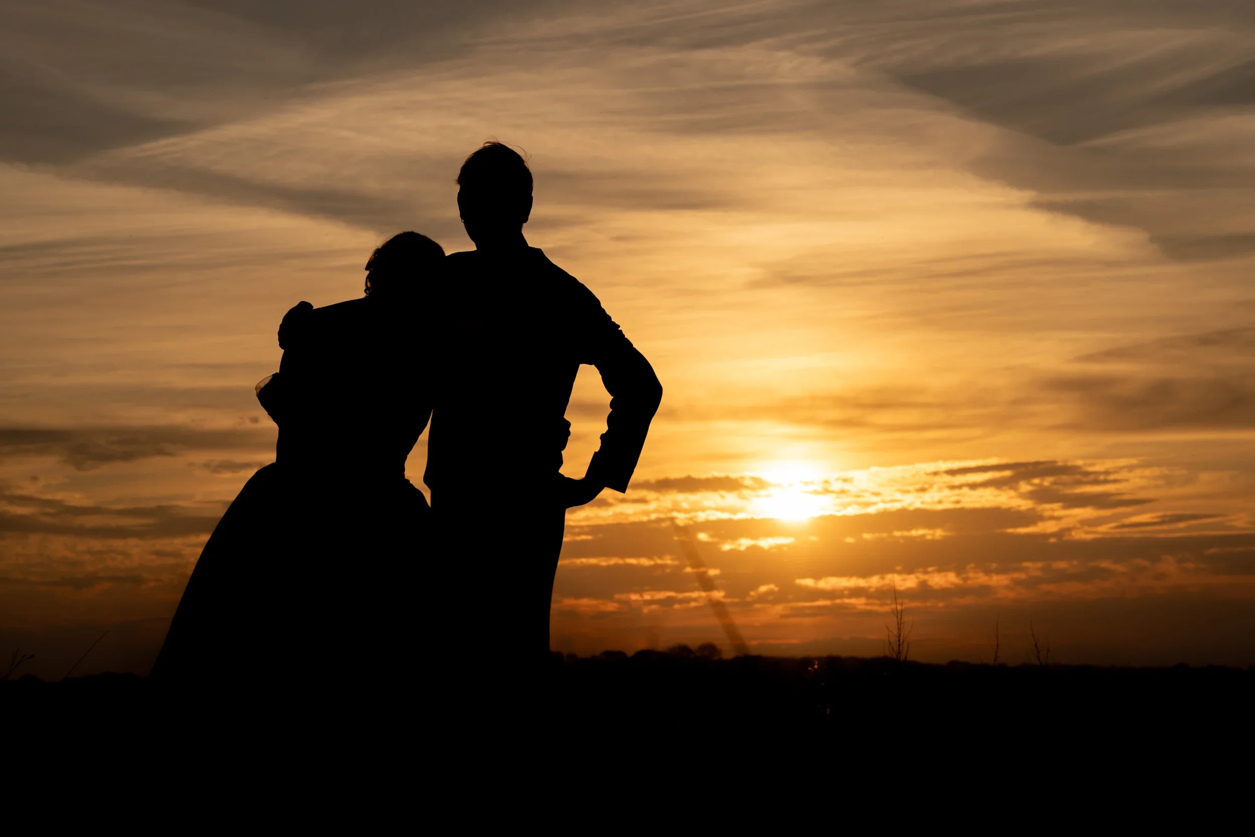 Silhouettes of two people, one adult and one child, against a sunset sky with clouds.