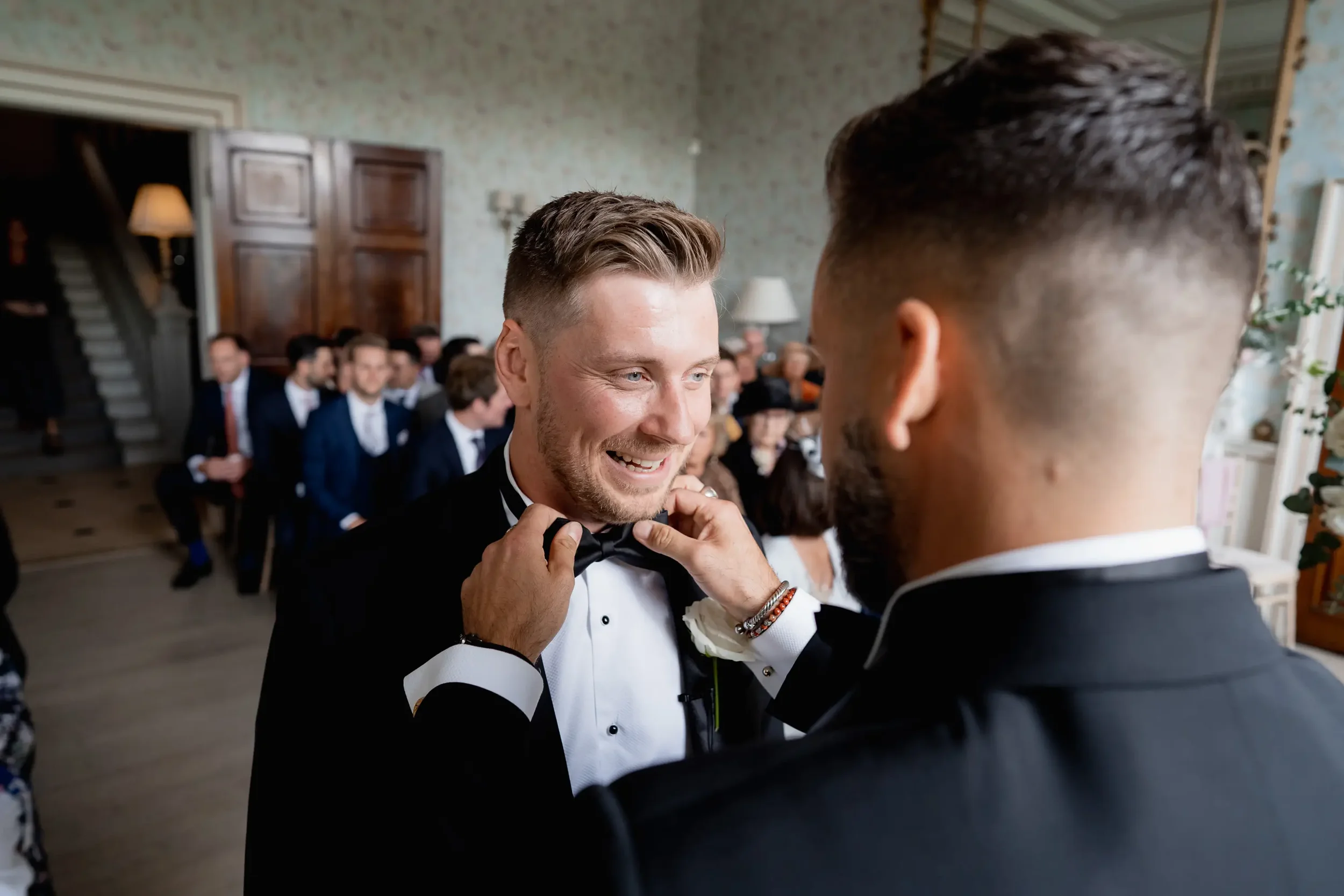 A groom adjusting his bow tie while smiling at another man during a wedding ceremony, with guests seated in the background.