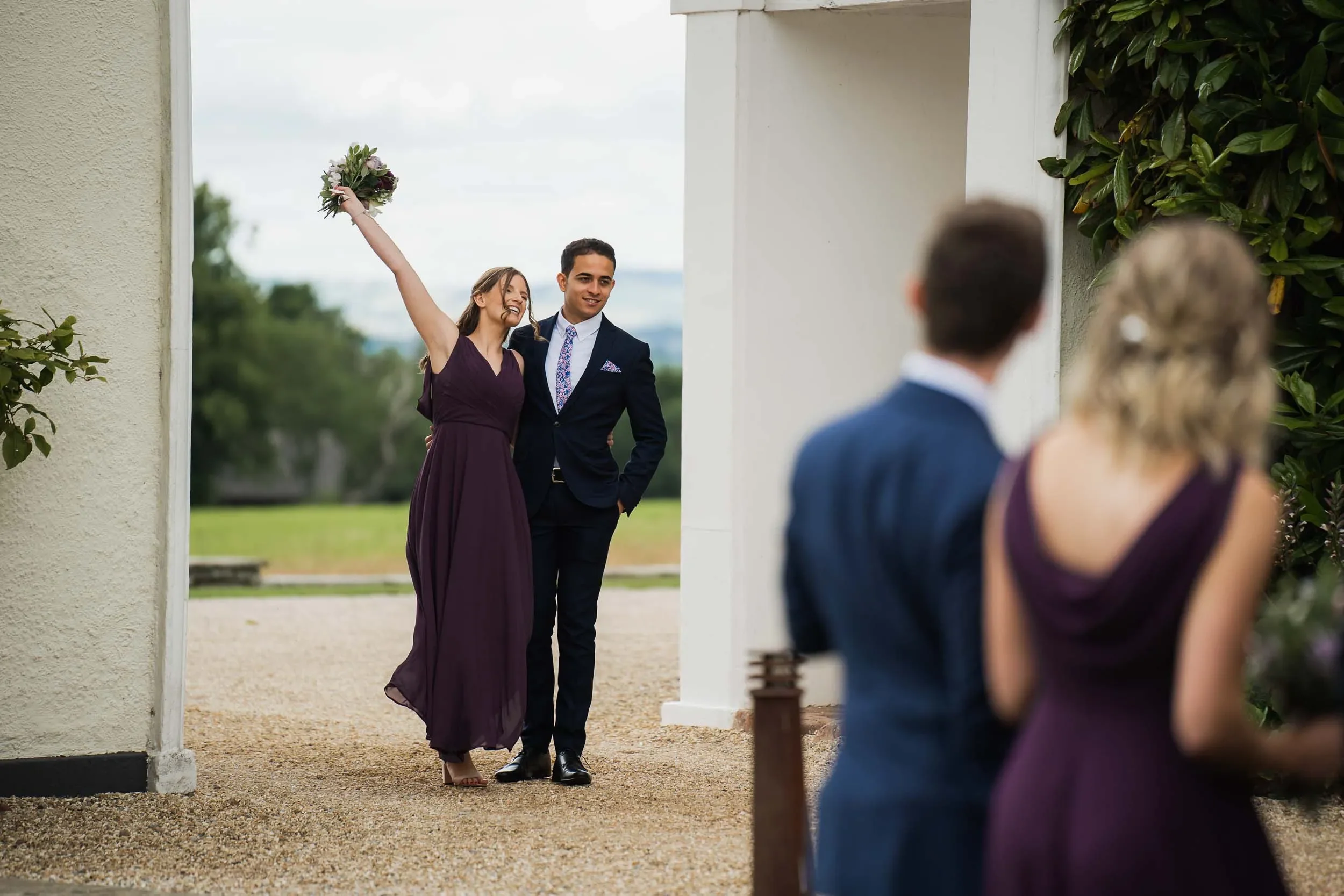 A woman in a purple dress holding a bouquet, celebrating as she walks with a man in a dark suit, outside near a white building with greenery, while a couple in the foreground watches.