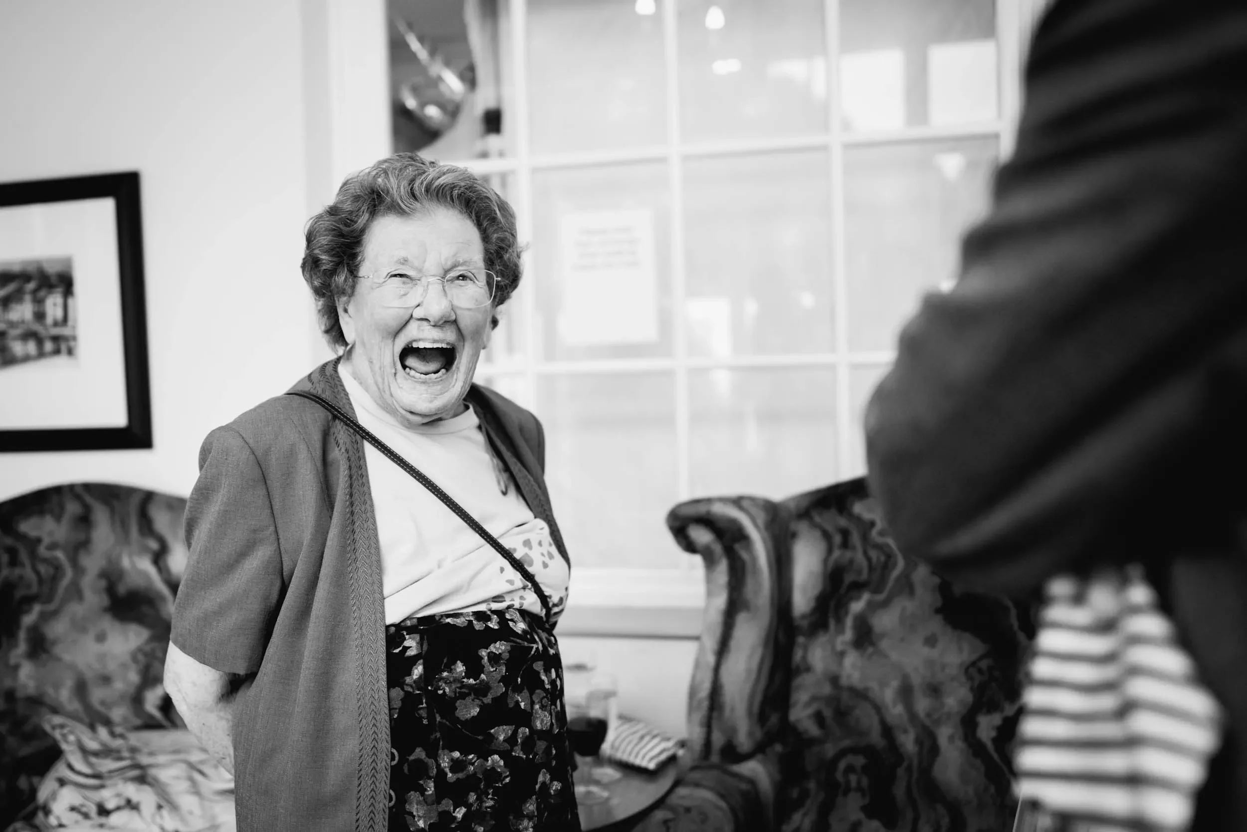 An elderly woman with glasses, smiling and laughing joyfully, standing in a room with a patterned couch and a window in the background.