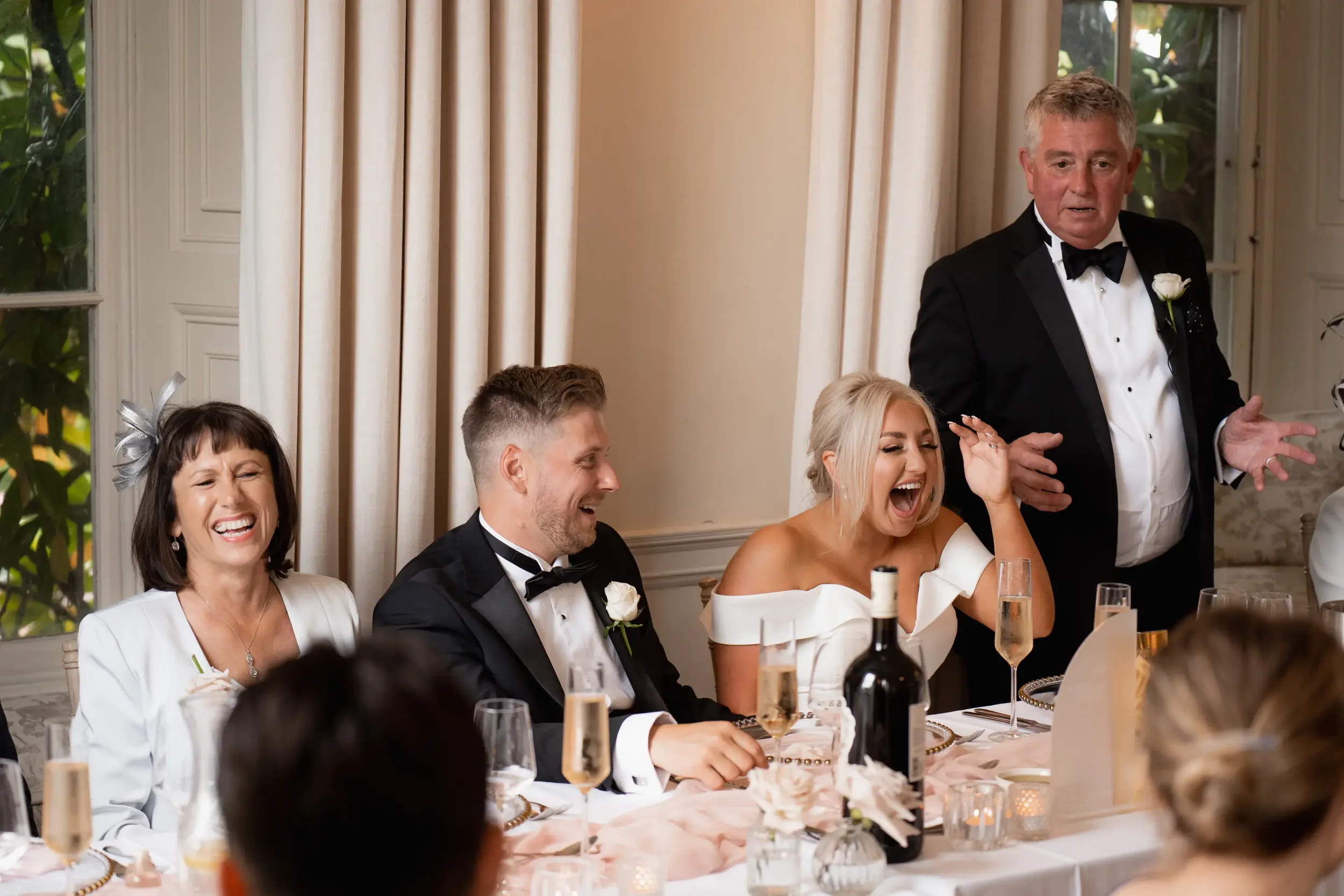 People seated at a wedding reception table, laughing and smiling, with a man standing and speaking, in a well-lit room with large windows and curtains.