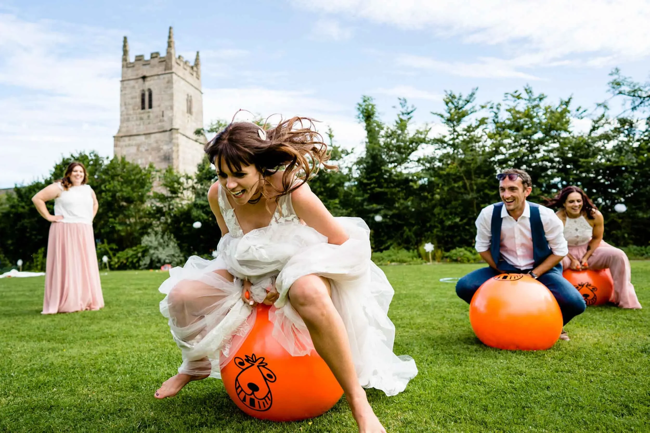 Group of people playing a game on grass with a historic tower in the background. One woman in a white dress is riding on an orange ball, while others watch and smile.