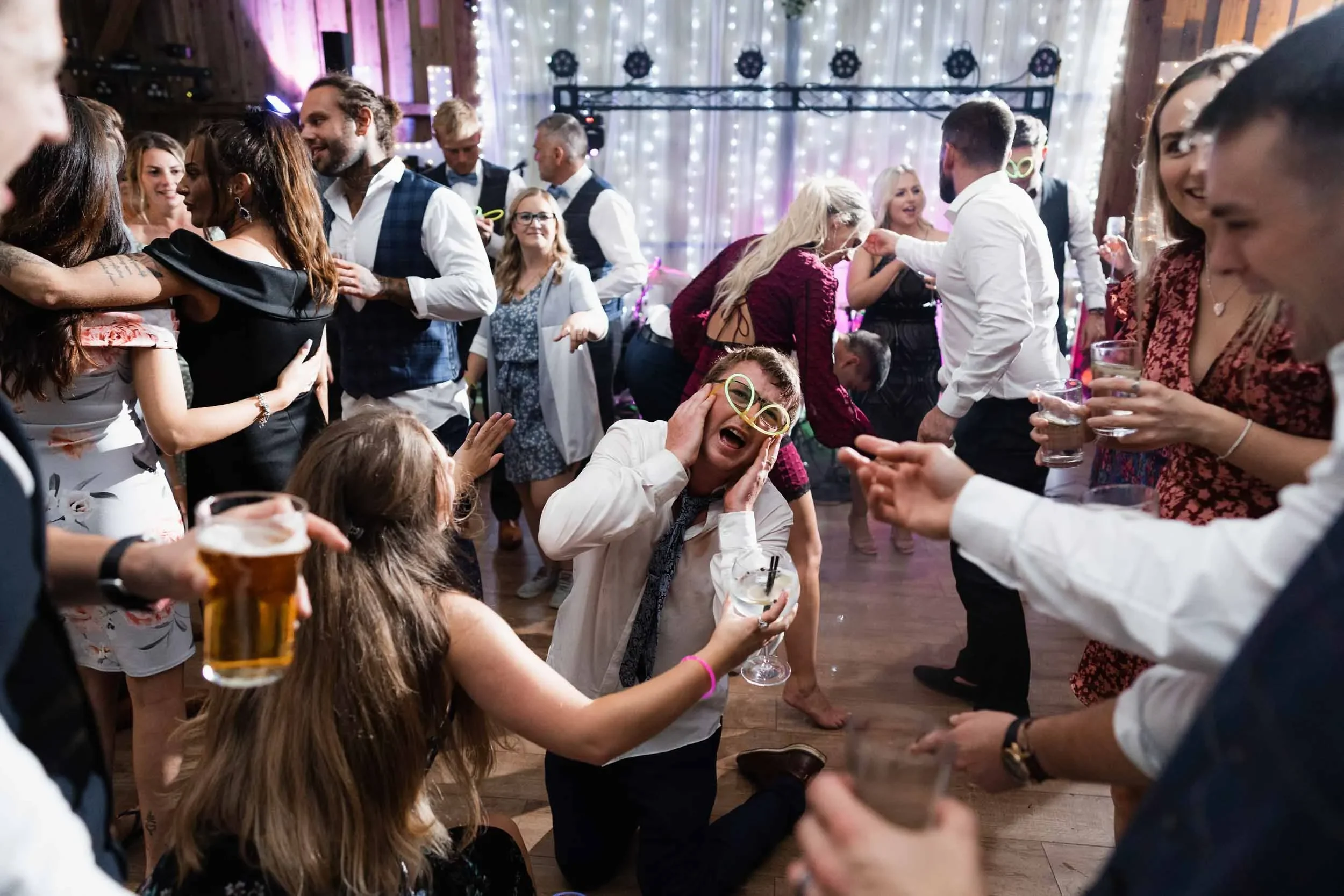 People dancing and celebrating at a party with colorful lights and a stage in the background.