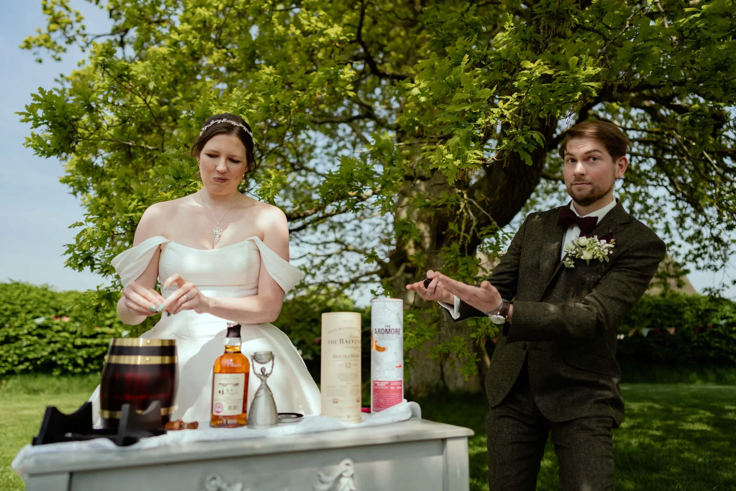 A woman in a white wedding dress and a man in a suit outside under a large tree, standing near a table with bottles of alcohol and cups, with some items in her hands and the man gesturing with his hands.