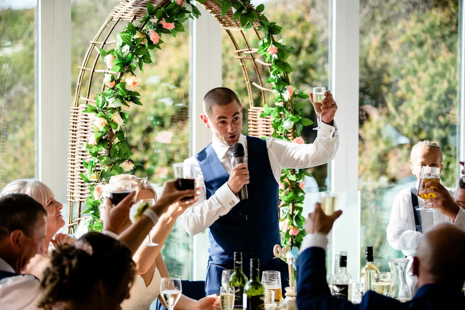 A man in a blue vest and white shirt giving a speech with a microphone, holding a glass of champagne, in a decorated setting with guests raising glasses at a celebration.
