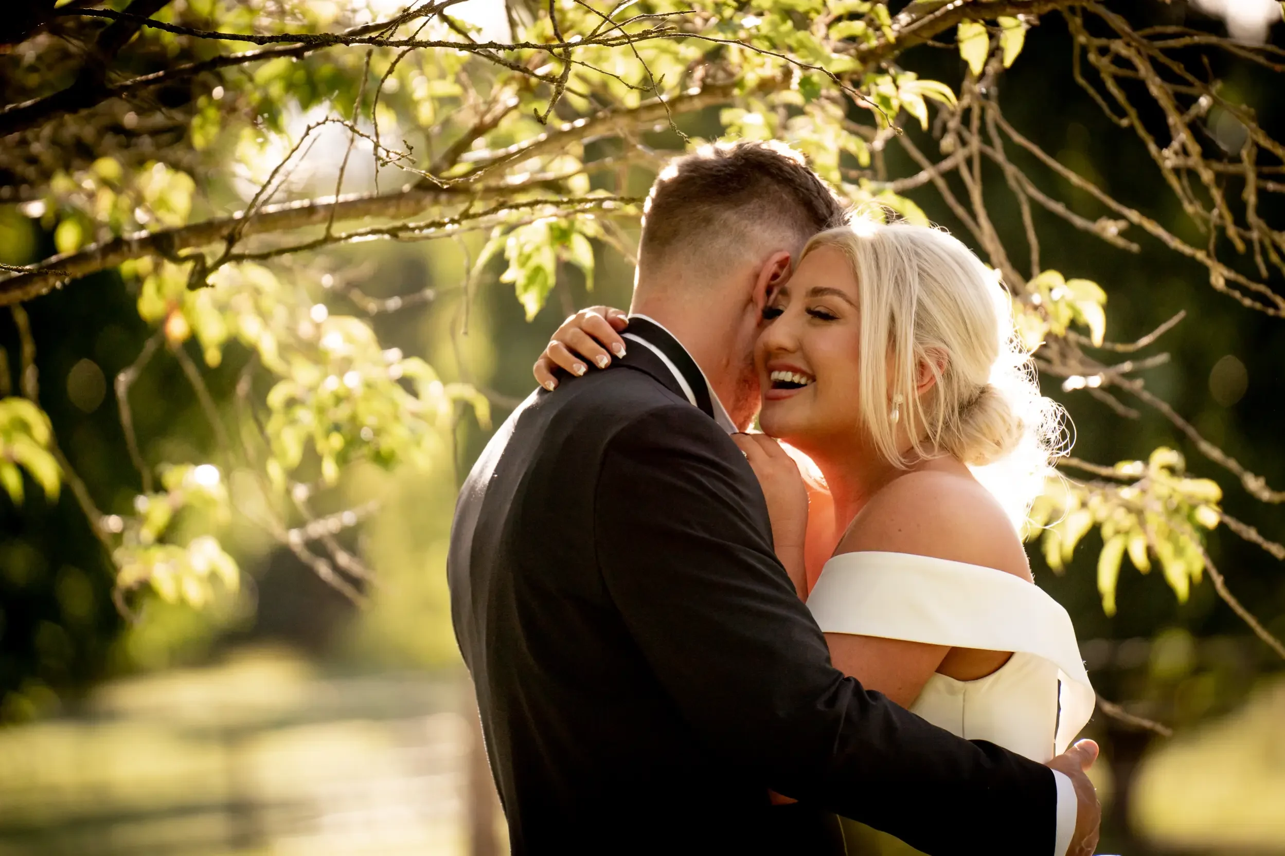 A newlywed couple embraces outdoors during sunset, the bride smiling with eyes closed, wearing a white off-shoulder dress and pearl earrings, the groom in a black tuxedo with a white shirt, with lush green trees and sunlight in the background.