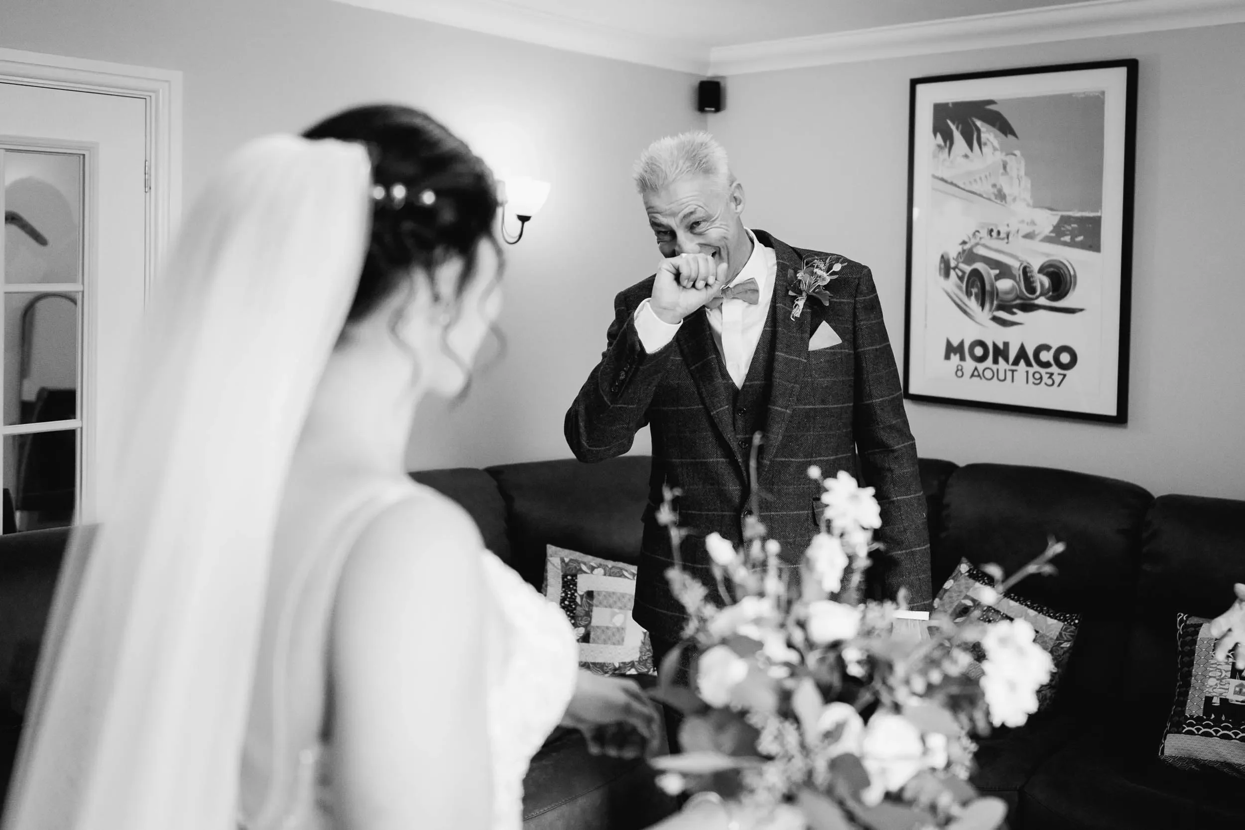 A black-and-white photo of a wedding scene with a bride in a veil and a man in a plaid suit, likely the groom, sharing a joyful moment inside a living room decorated with a Monaco poster and floral arrangements.