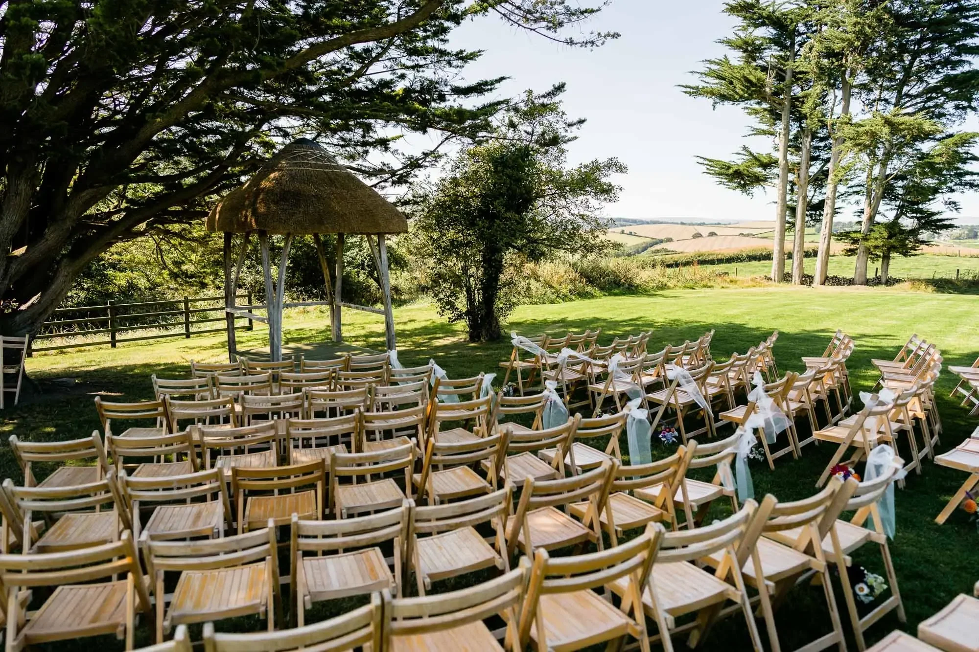 Outdoor wedding setup with wooden chairs arranged in rows, decorated with white ribbons and flowers, under a thatched gazebo in a grassy field surrounded by trees and open countryside.