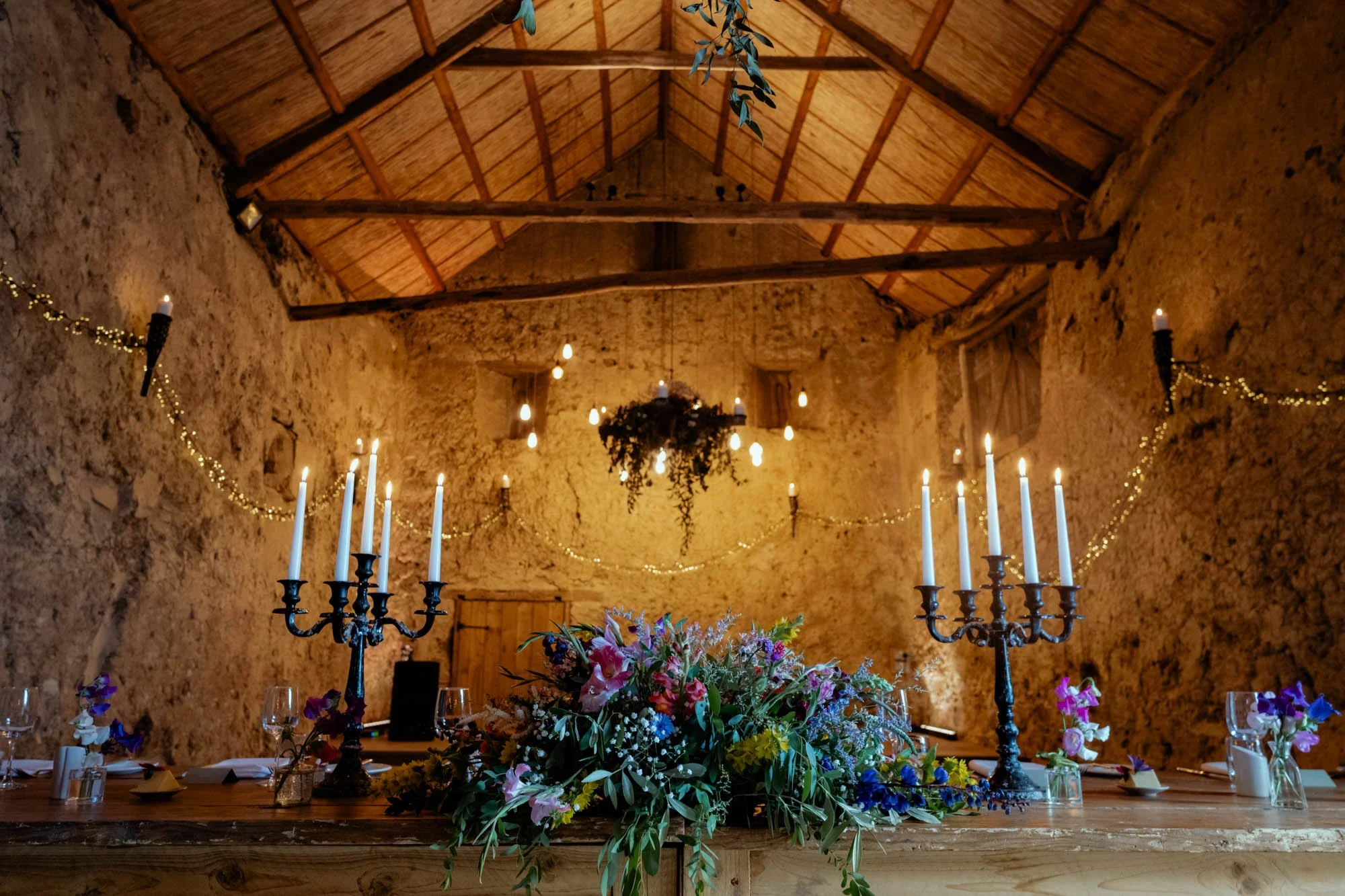 Wedding reception setup with a large floral centerpiece, candelabras with lit candles, and decorated with fairy lights inside a rustic stone barn with wooden ceiling beams.