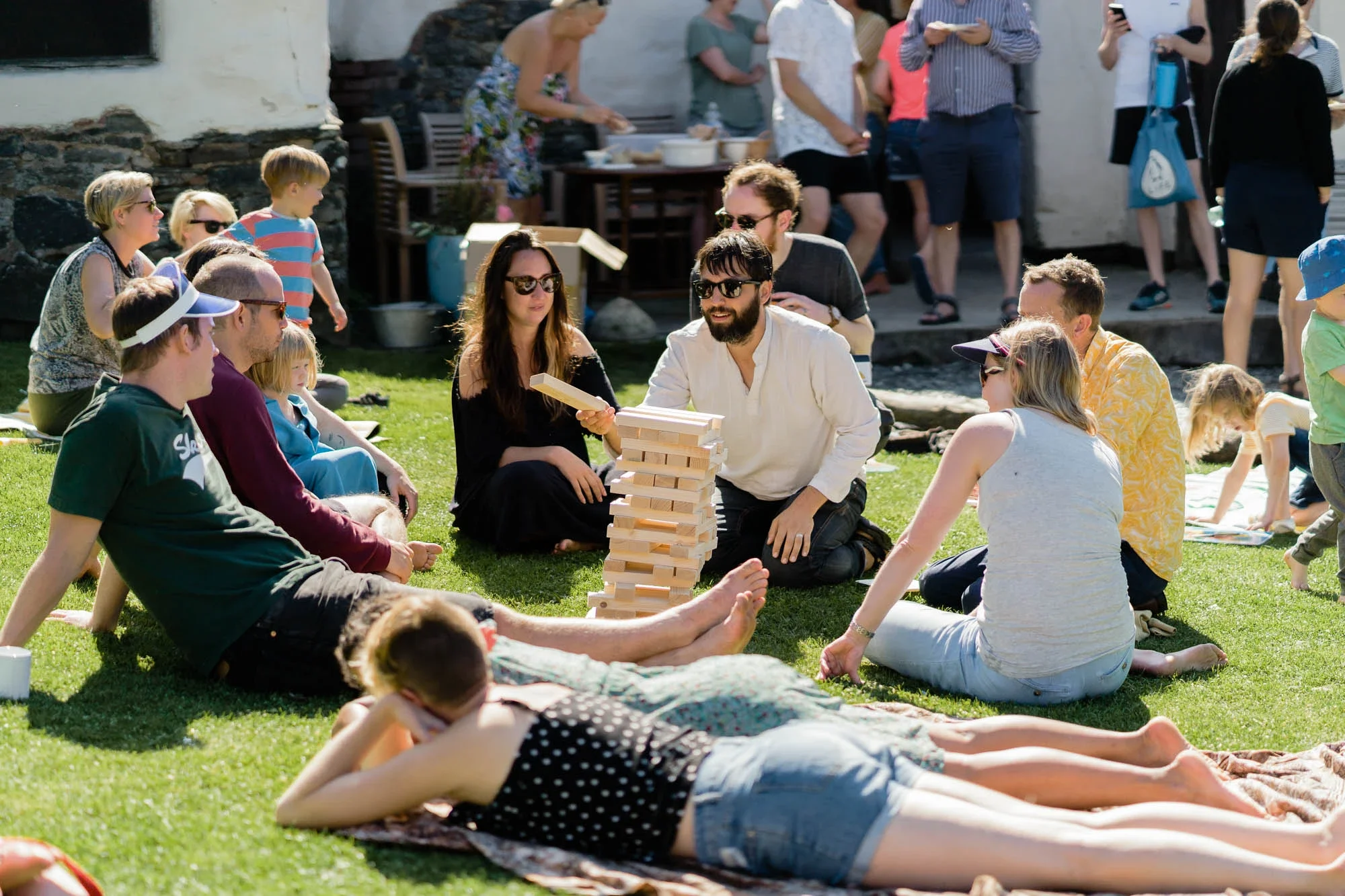 People gathered outside on a sunny day playing giant Jenga and relaxing on the grass, with others in the background socializing and enjoying food.