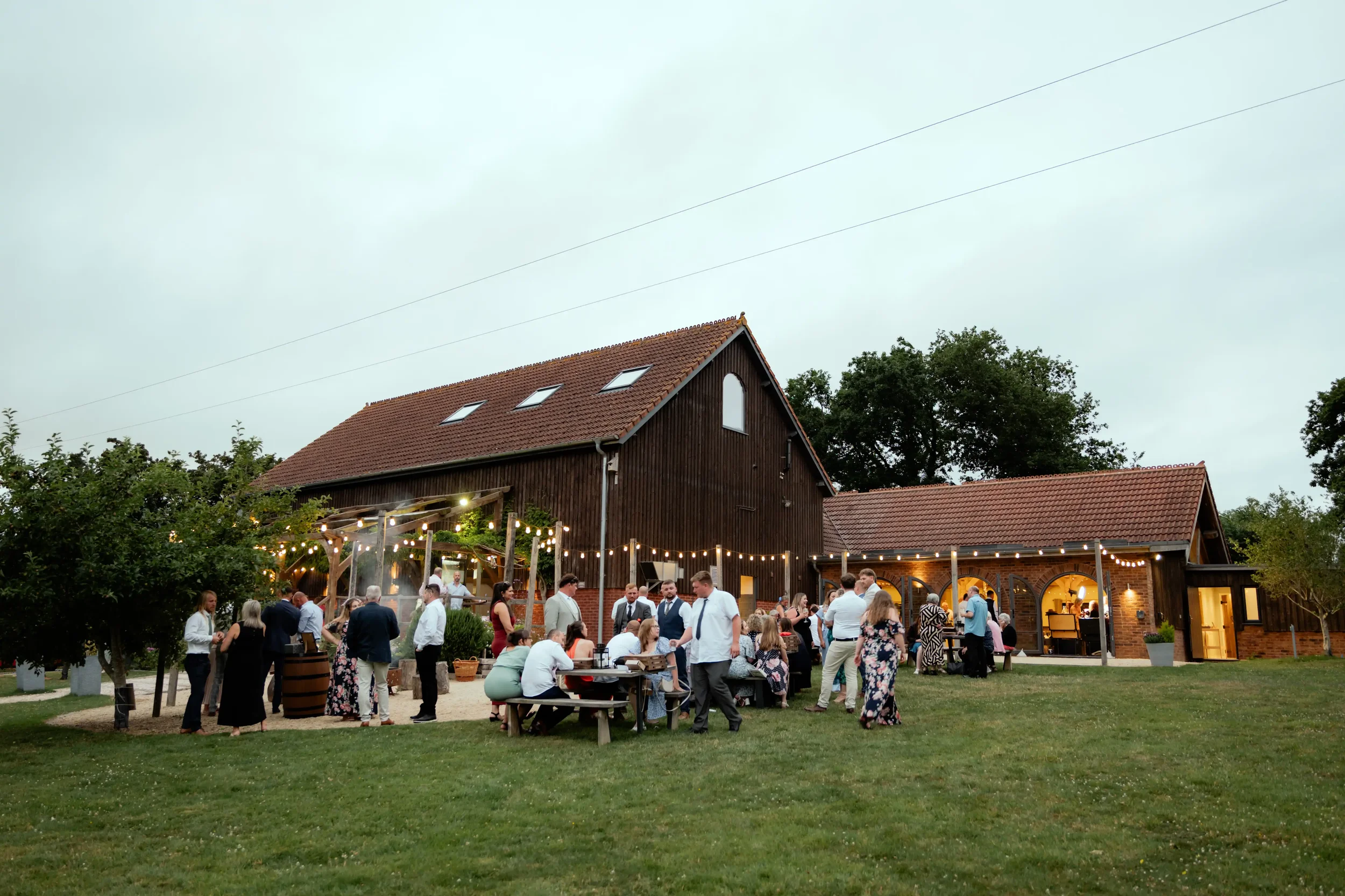 People gathered outdoors at evening wedding reception with string lights, seated and standing around tables on grassy lawn near rustic barn-style building.