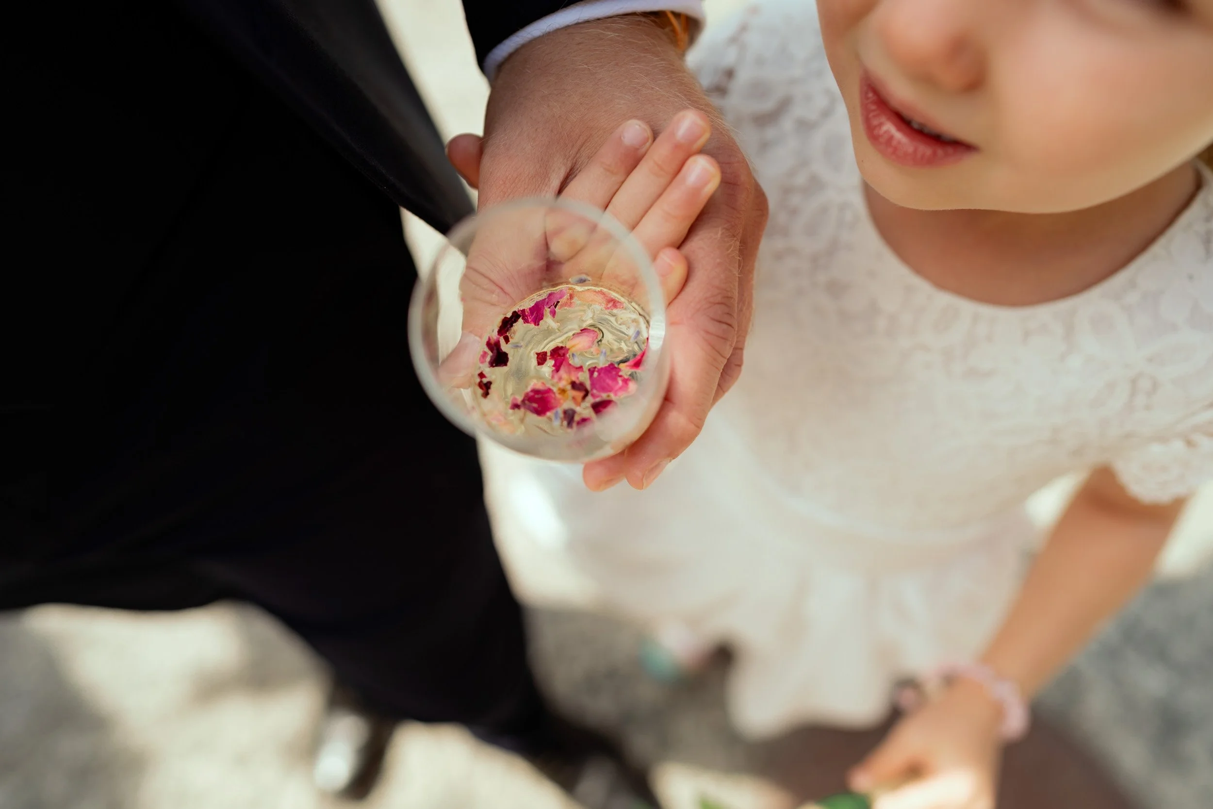 A young girl in a white lace dress and a pink bracelet is holding a glass of champagne with pink flower petals floating on top, as a man in a black suit touches her head.