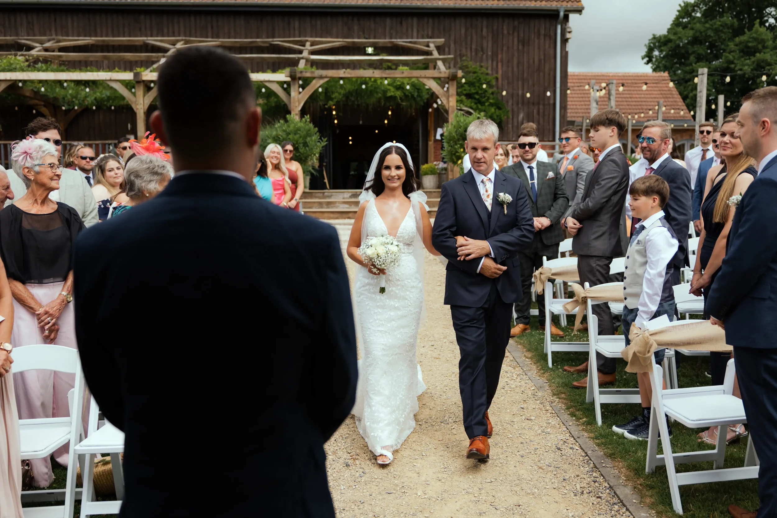 Bride walking down the aisle with her father at a wedding ceremony outdoors, surrounded by guests seated on both sides.