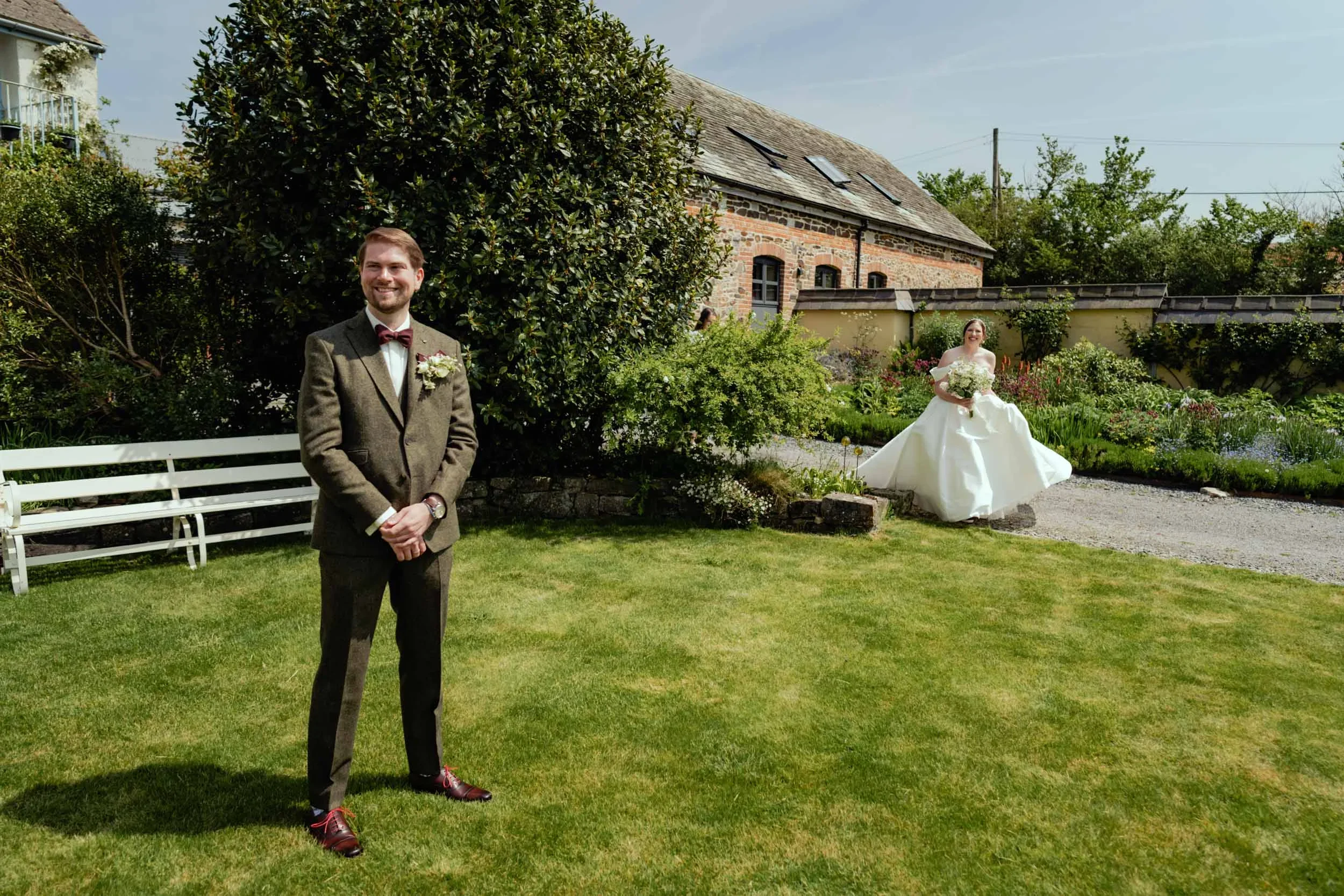 A groom standing on a lush green lawn in a garden, smiling, wearing a brown suit with a bow tie, and a bride in a white wedding gown holding a bouquet, standing behind him near a garden path with colorful flowers, trees, a brick house, and a bench.